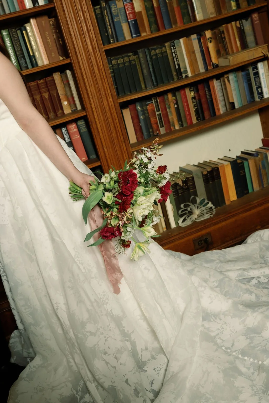A woman in a white wedding dress holding a bouquet of red, white, and green flowers, standing in front of a wooden bookshelf filled with books.