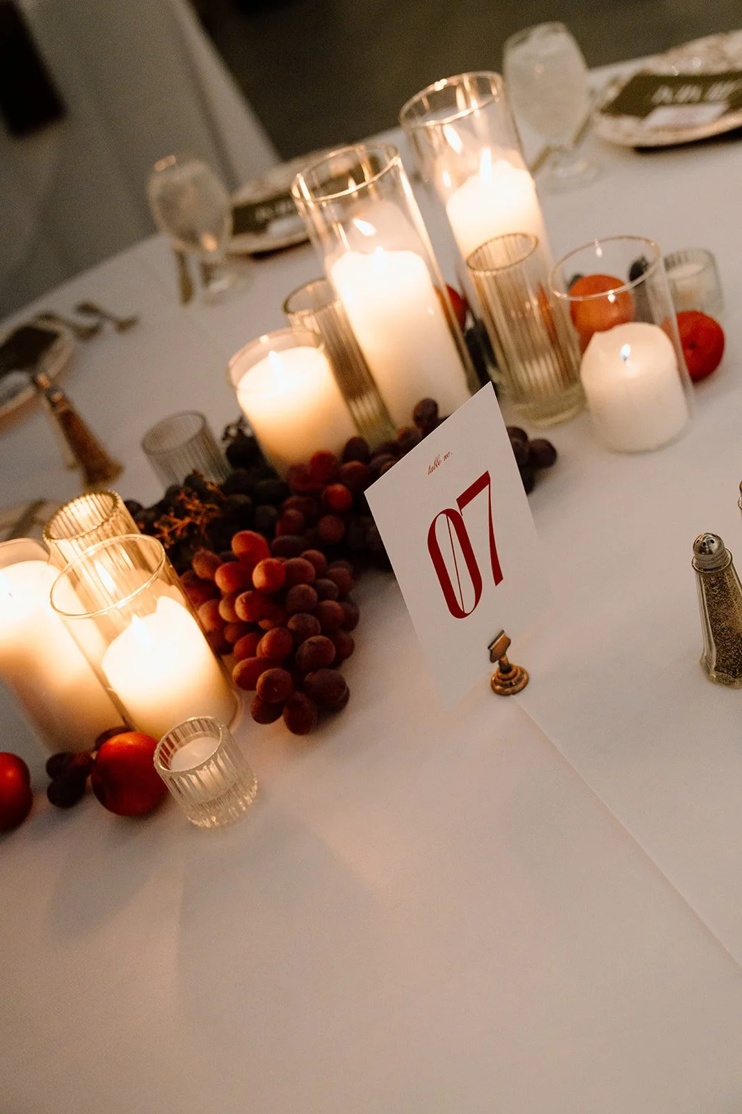 Table decorated with white candles in glass holders, bunches of red grapes, small apples, and a table number card labeled '07' at a formal event.