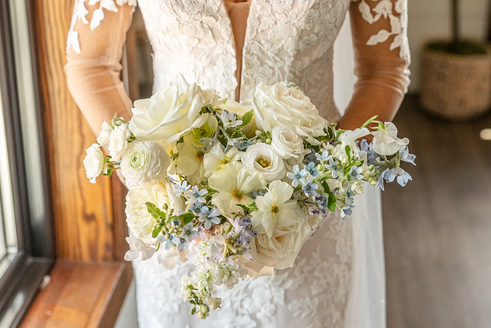 A bride in a lace wedding dress holding a bouquet of white flowers with some light blue accents, near a window with wooden framing.