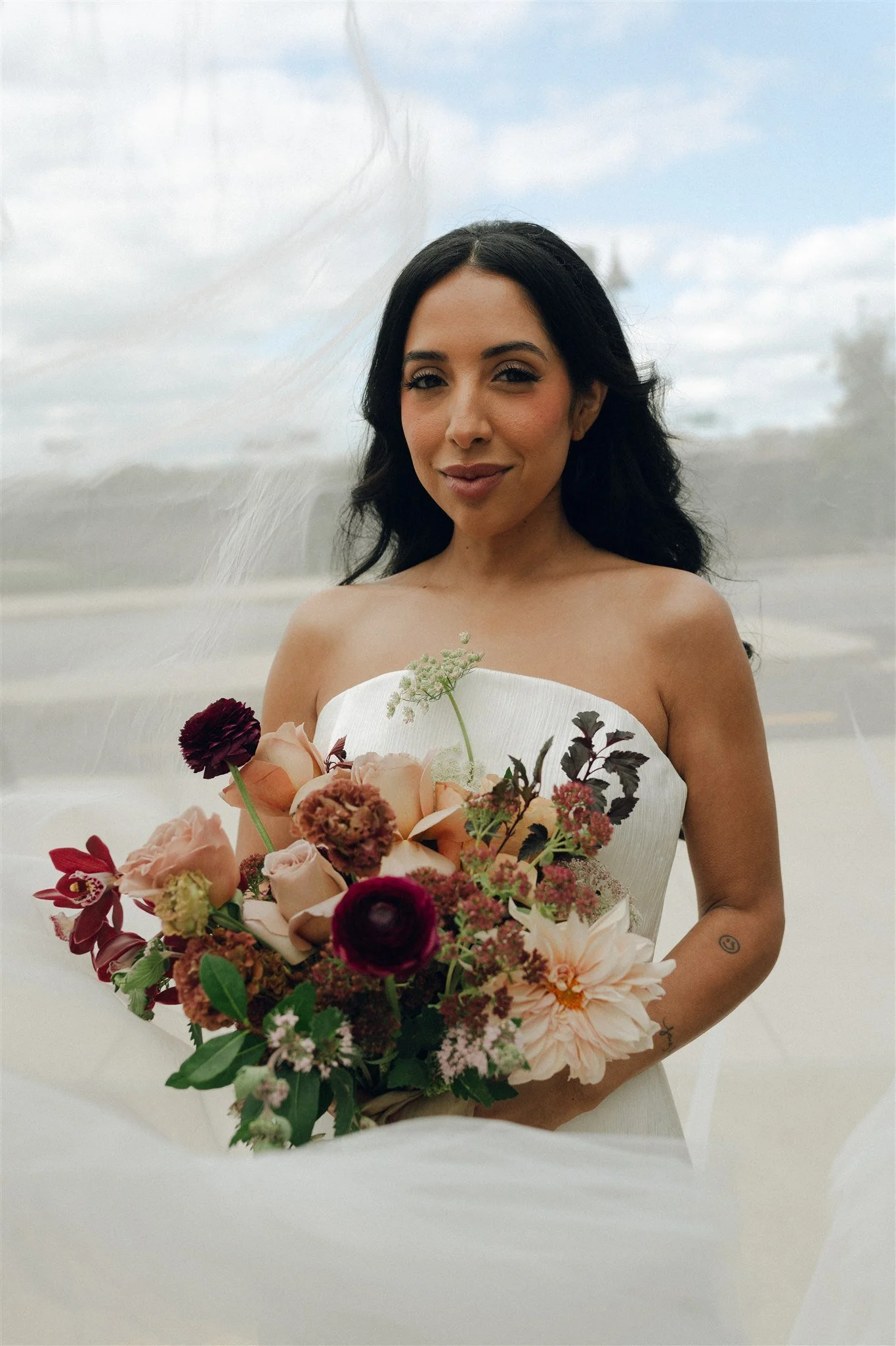 Woman in a strapless white dress holding a bouquet of pink, red, and peach flowers, standing outdoors against a cloudy sky.
