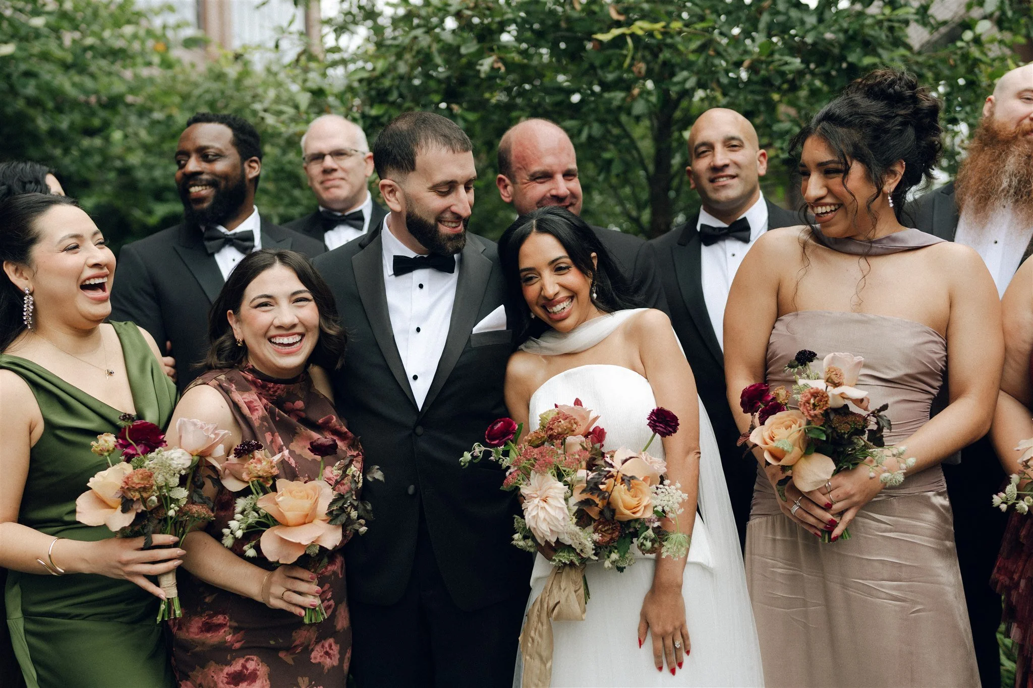 Wedding group photo with the bride, groom, bridesmaids, and groomsmen outdoors, all smiling and holding bouquets, dressed in formal attire.