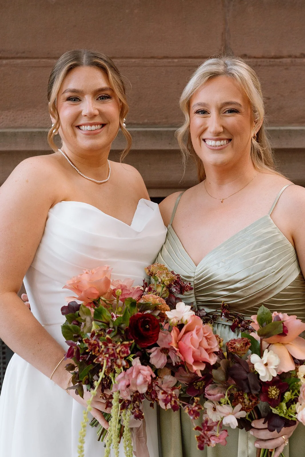 Two women smiling at a wedding, one in a white wedding dress and the other in a green dress, holding colorful bouquets.
