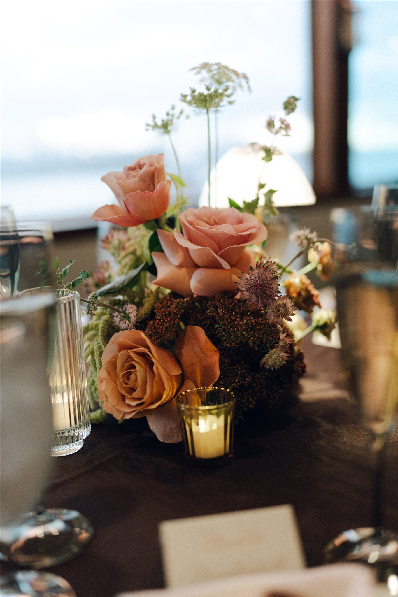 Close-up of a floral centerpiece with pink and peach roses, purple and green accents, on a dark table with gold and clear glass candle holders in a softly lit setting.