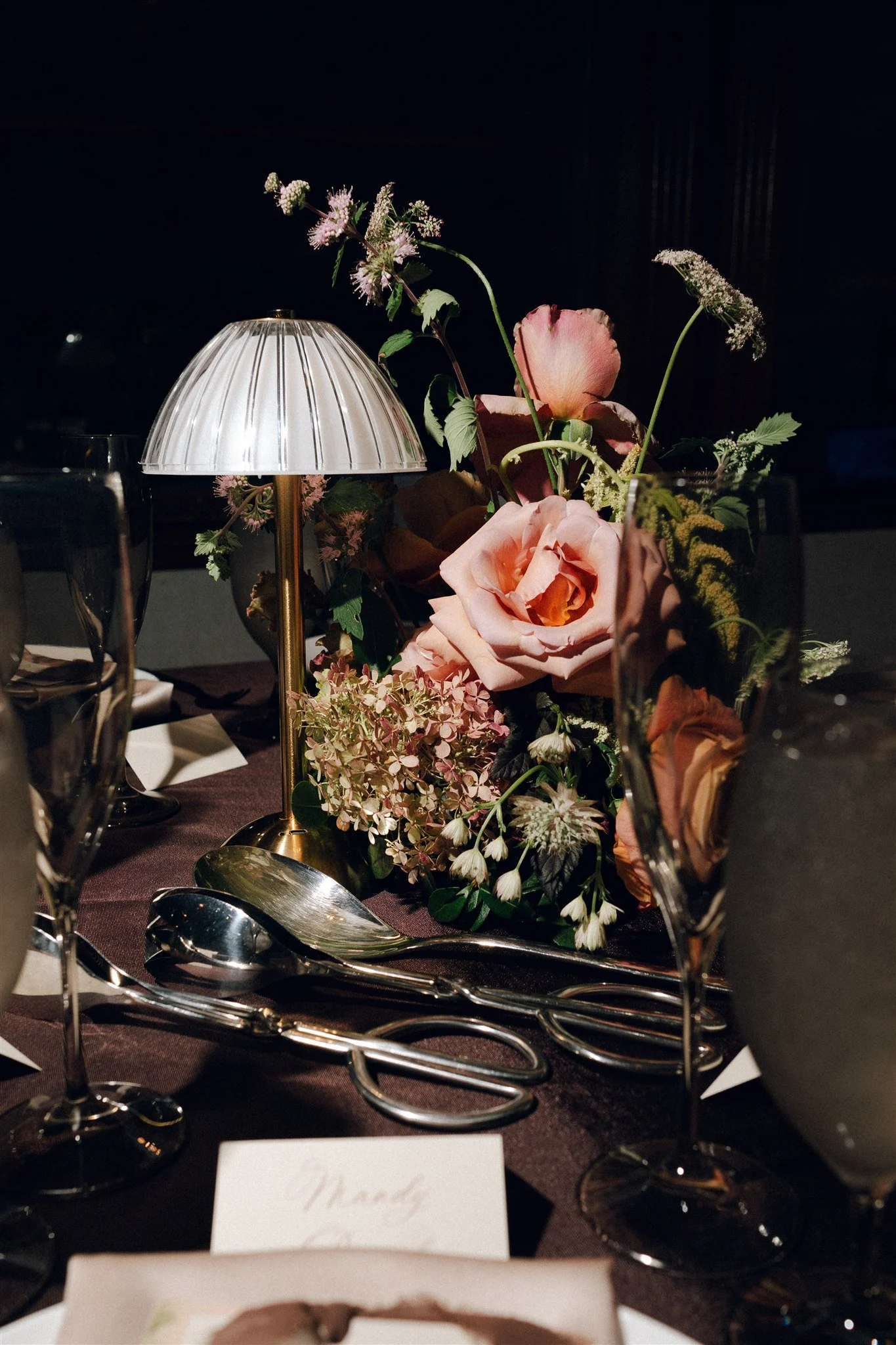 A dinner table with a floral centerpiece, a small white table lamp, and silverware, set for a formal event.