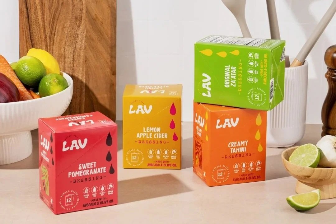 Three colorful boxes of Lav salad dressing on a kitchen counter, with bowls of limes and other fruits nearby. Photography for Dress Lav. The Apricots Media. Social Media Agency for CPG, DTC and Hospitality brands.