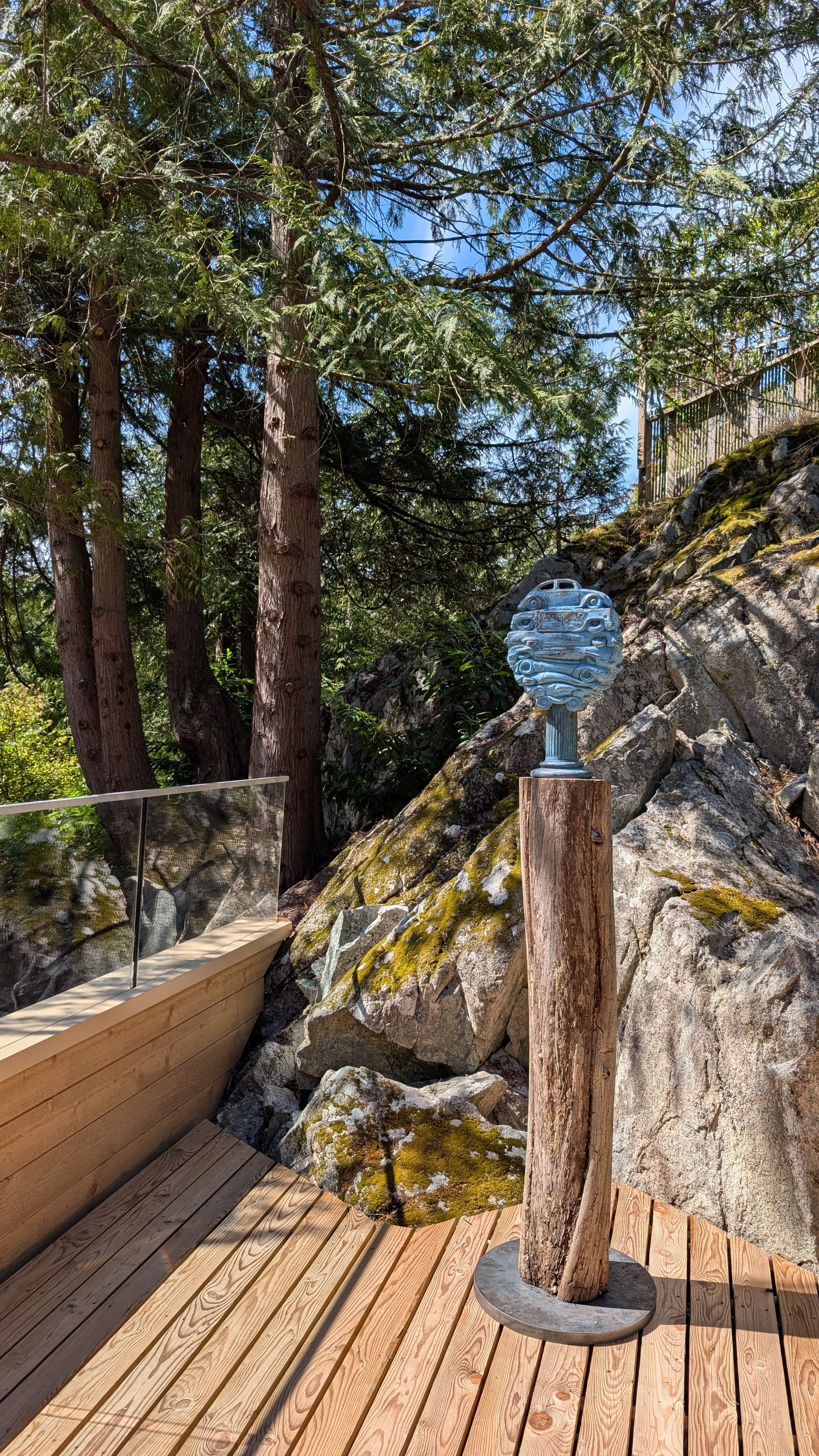 An outdoor scene with tall trees, a rocky hillside, and a wooden deck. There is a sculpture of a blue vintage car mounted on a wooden post and a glass railing on the deck.