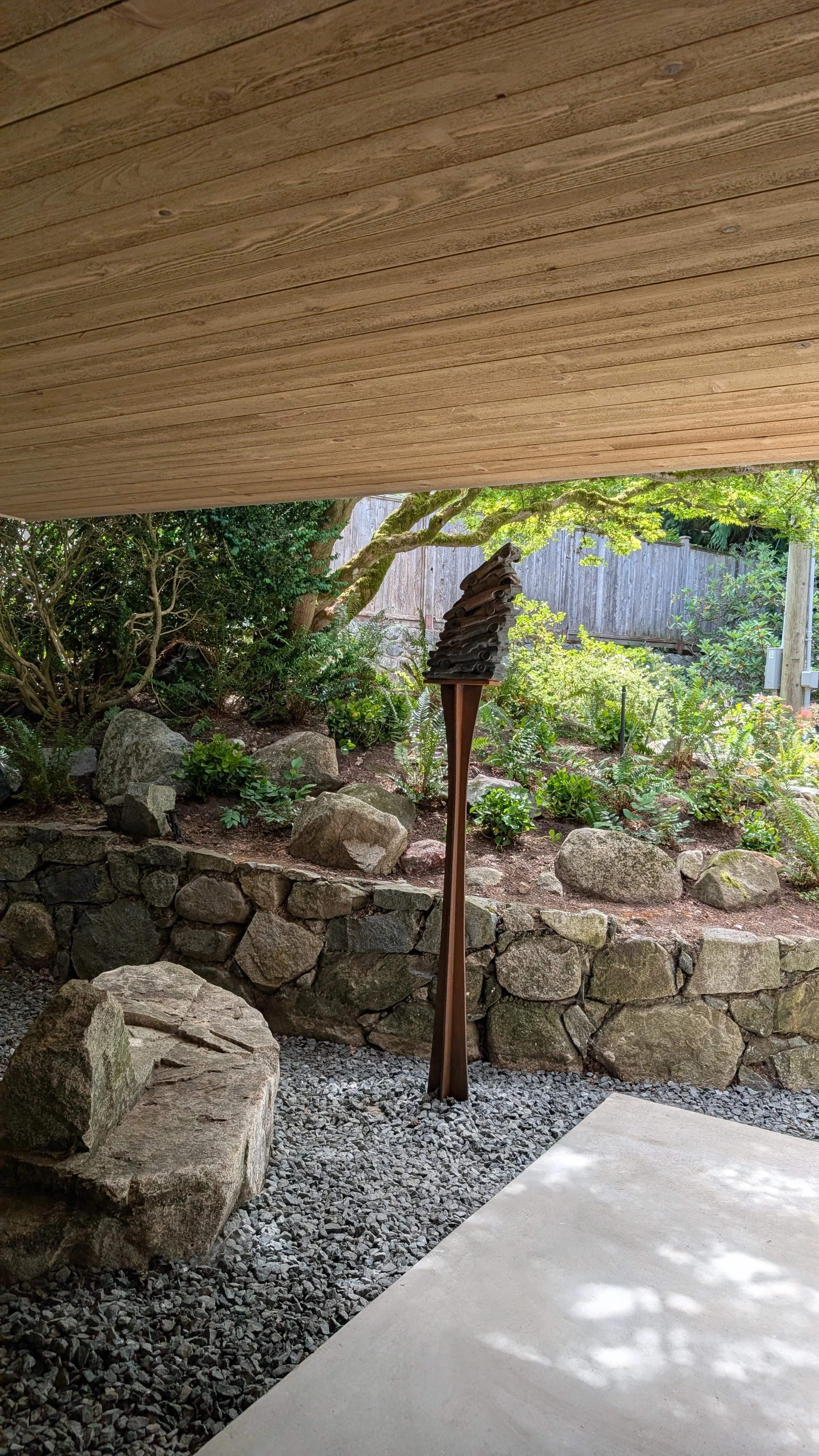 View of a small rock garden with a variety of greenery, trees, and a wooden fence in the background, seen from underneath a wooden overhang.