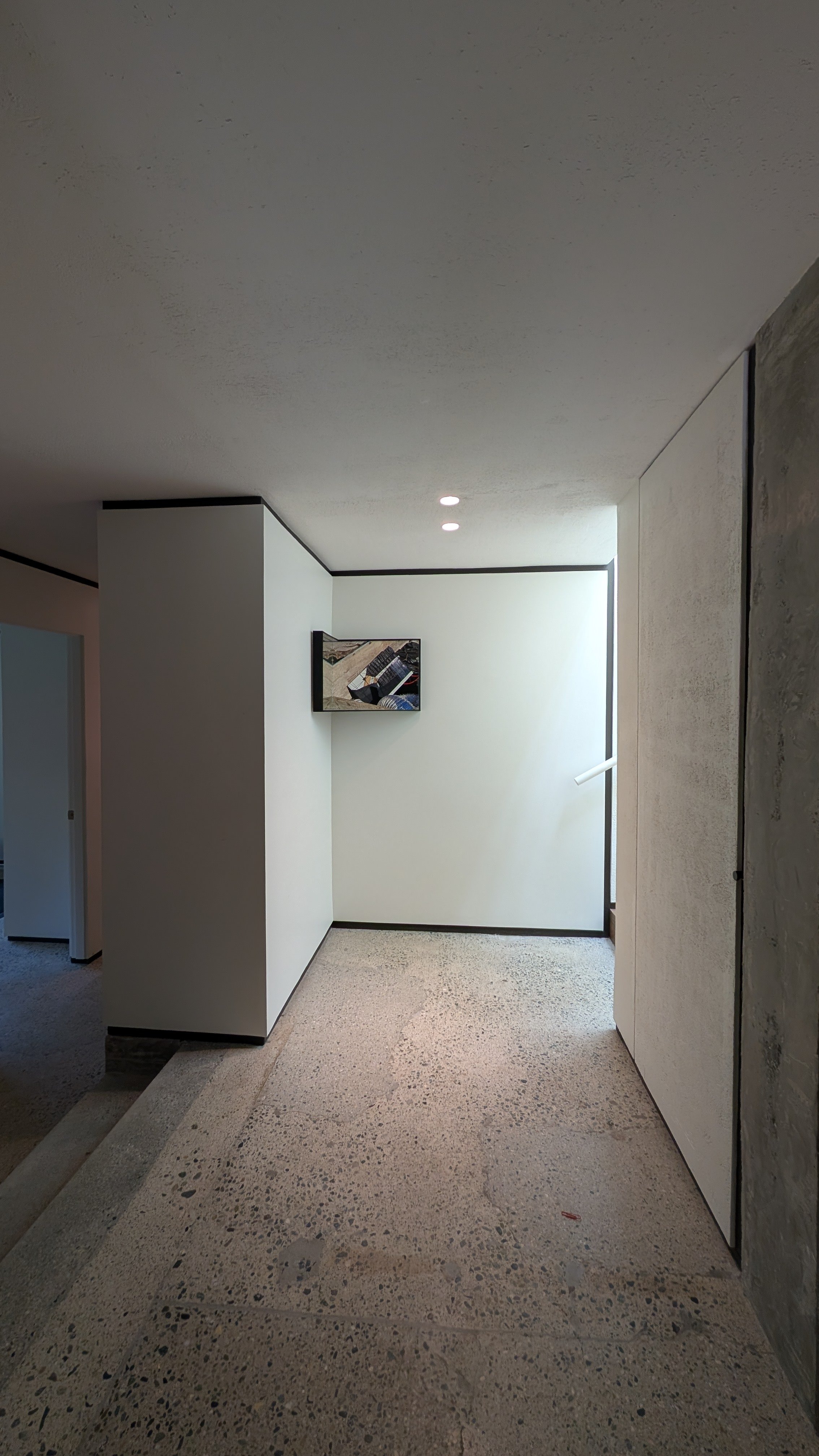 A minimalist modern interior space with light-colored terrazzo flooring, white walls, and a gray textured wall on the right. There is a small framed artwork on the back wall and recessed ceiling lights.