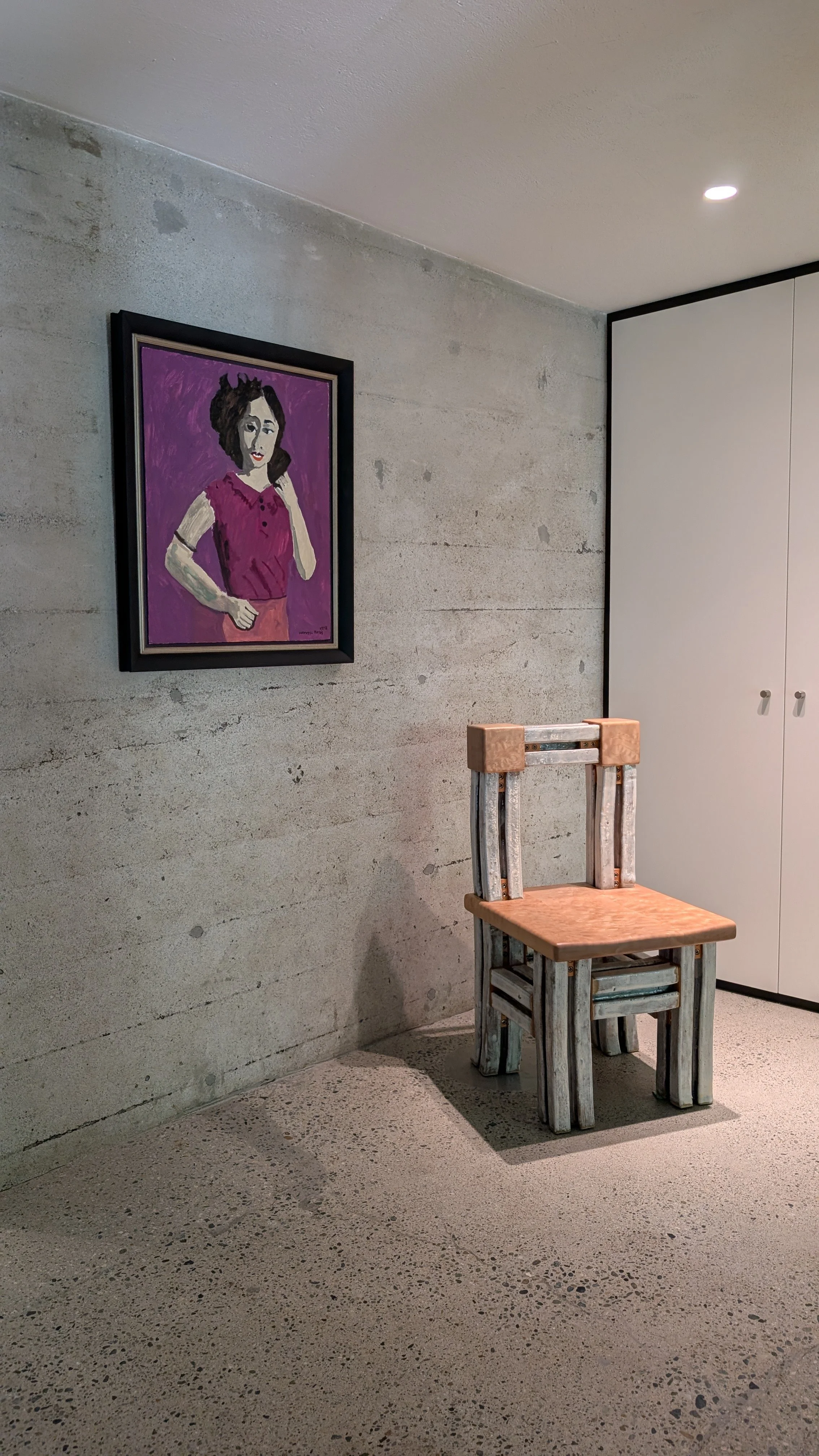 A room with a concrete wall, a framed portrait of a woman wearing a pink top, a wooden chair with a distressed painted finish, and a white cabinet in the background, with a small ceiling light.