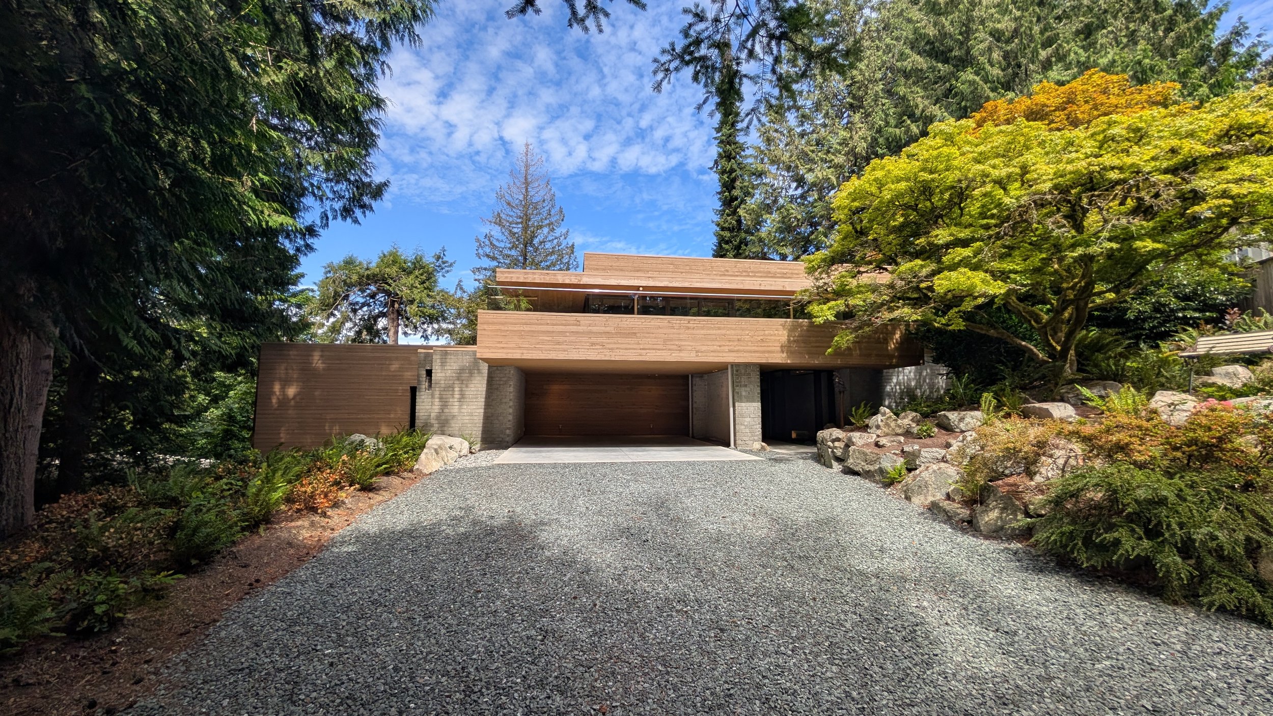 Modern wooden house with large windows, surrounded by lush green trees and plants, on a gravel driveway under a partly cloudy sky.