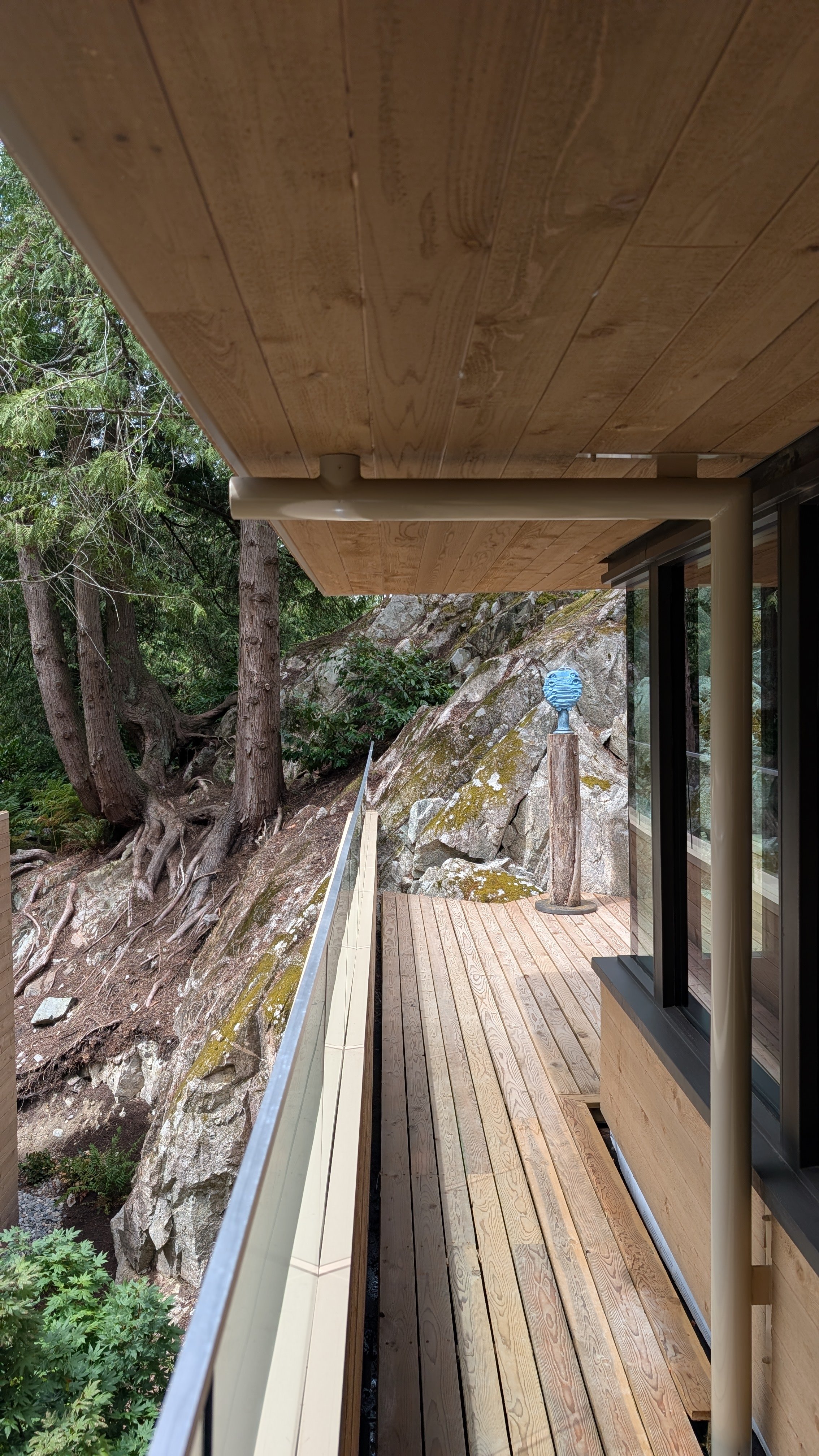 A wooden deck extending from a modern house built into a rocky hillside, with a view of trees and rocks outside, and a sculpture on a stand in the background.