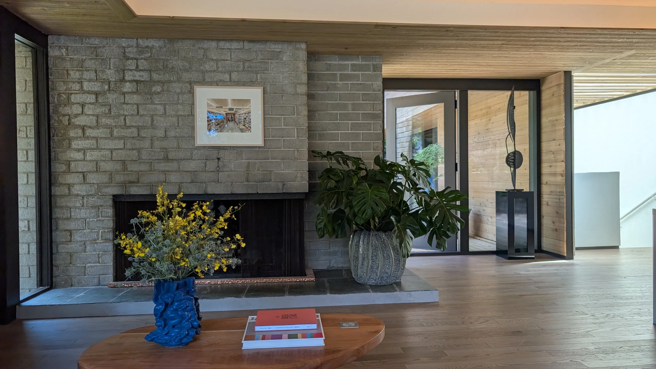 Interior of a modern house featuring a brick fireplace, large green potted plant, and framed artwork on the wall, with doorways leading to an outdoor patio.