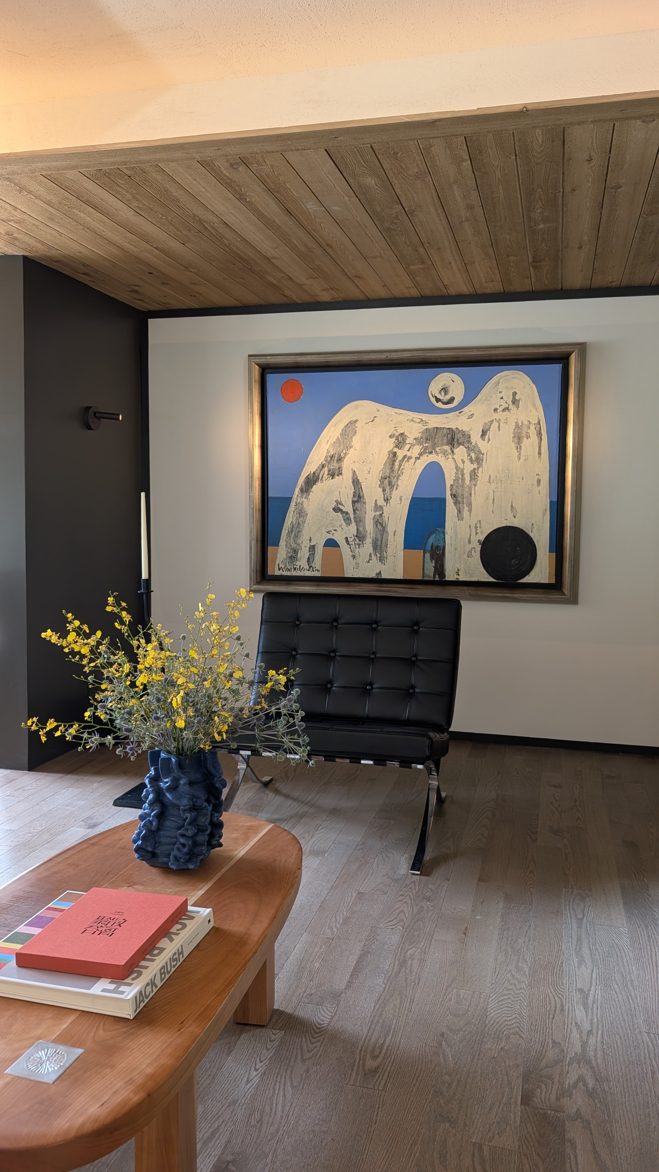 Living room corner with a black leather chair, a wooden coffee table with books, a vase of yellow flowers, a framed abstract painting of a horse, sea, and celestial bodies, and a wall-mounted light fixture.
