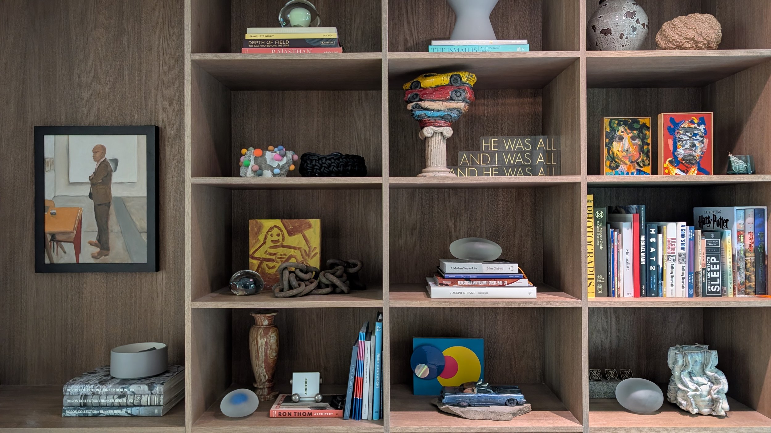 A wooden bookshelf with various decorative items, books, and artwork. The top shelf includes books and a light green sculpture, with a yellow toy car and stacked smaller cars in the middle. The second shelf features a painted portrait of a man, a sma