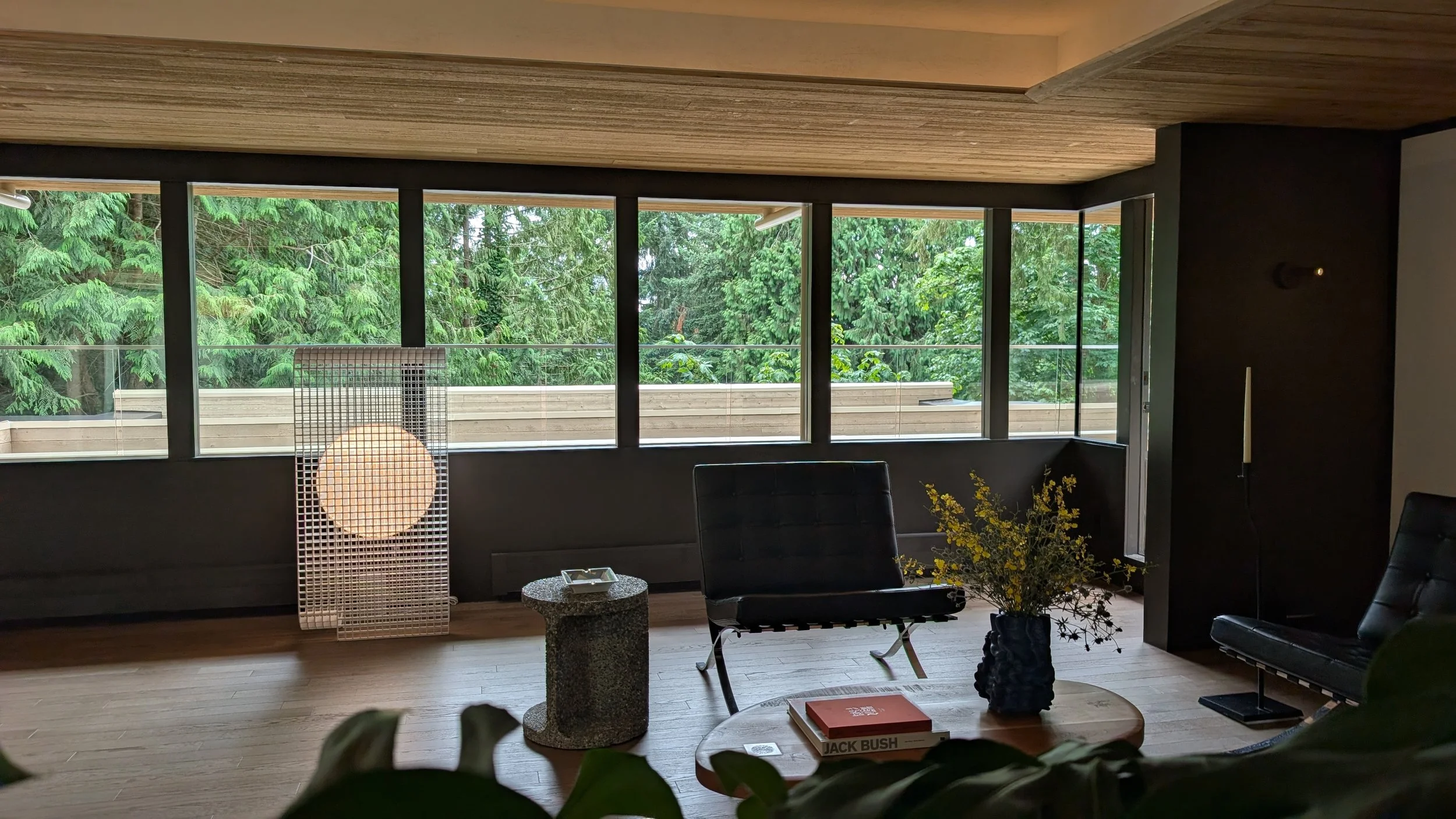Interior view of a room with large windows overlooking green trees, featuring modern furniture including black chairs, a round table with books, a decorative vase with yellow flowers, and a standing lamp with a spherical light. The ceiling has wooden