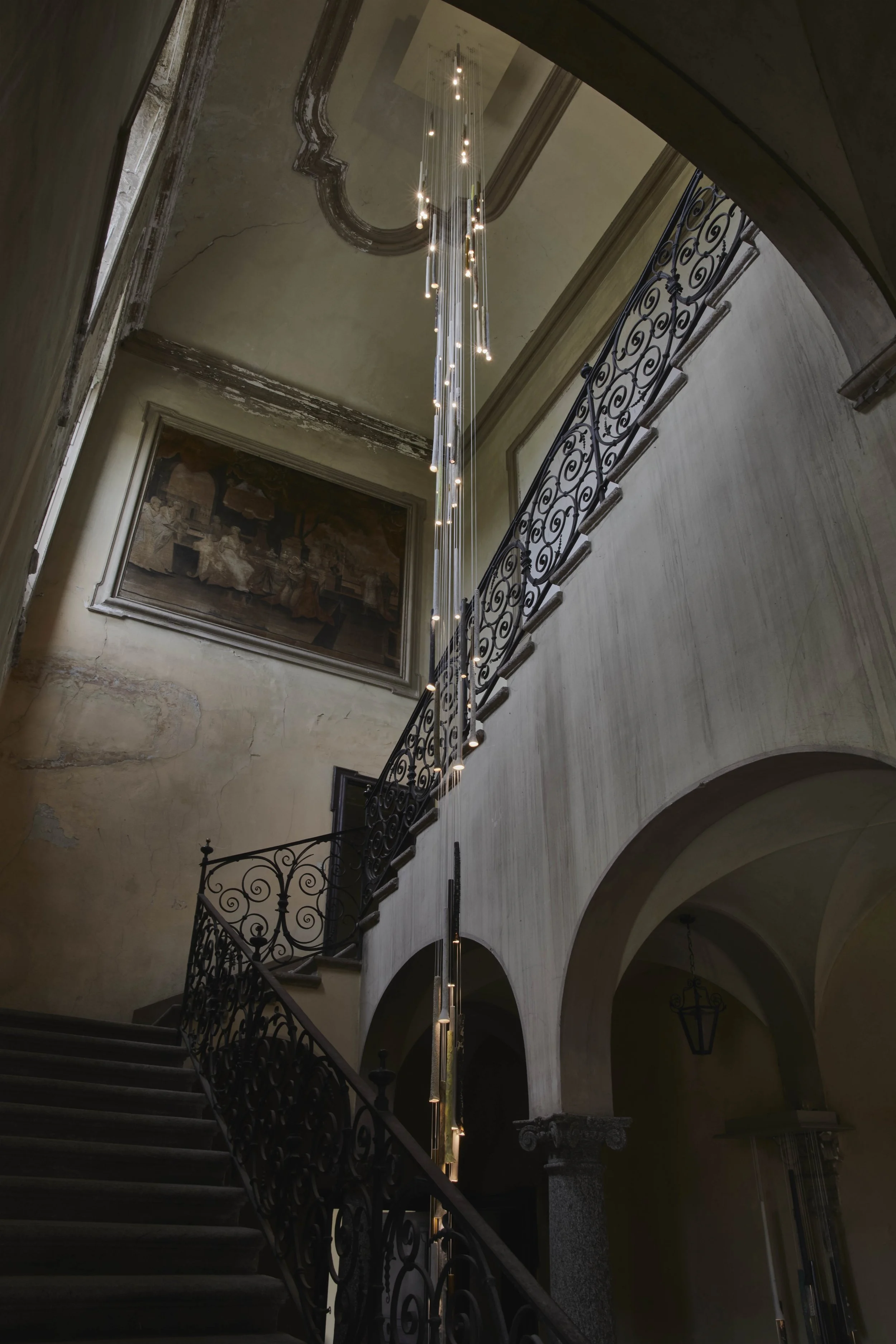 Interior view of a grand staircase in an old building with ornate iron railings, a large chandelier hanging from a high ceiling, and a classical painting on the wall.