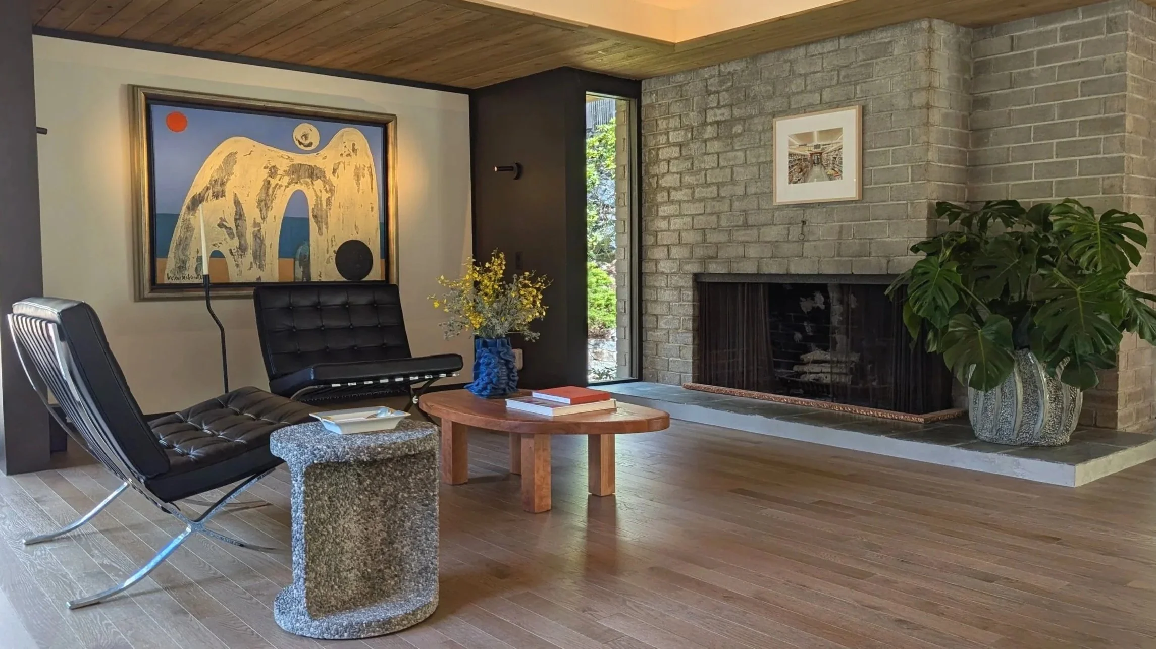 Living room with black leather lounge chairs, wood coffee table, fireplace, large houseplant, abstract artwork on wall, and a window showing greenery outside.