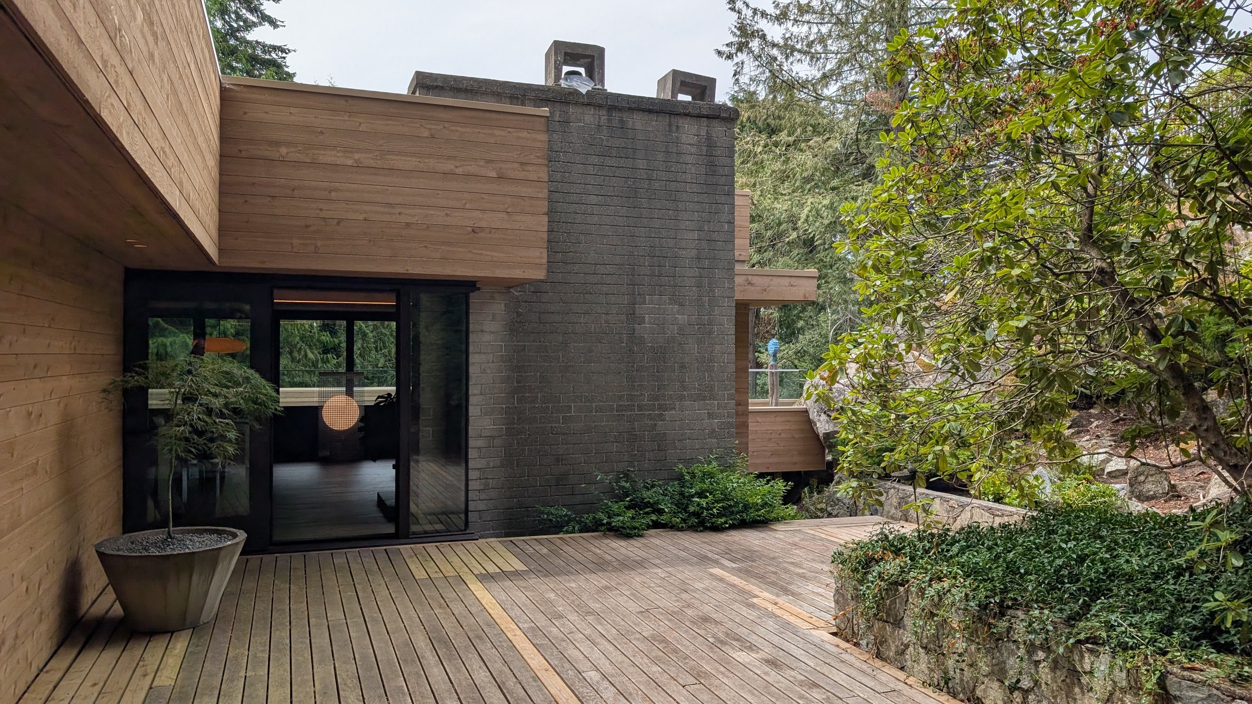 Modern house with wooden deck and large glass sliding doors, surrounded by trees and natural landscape.