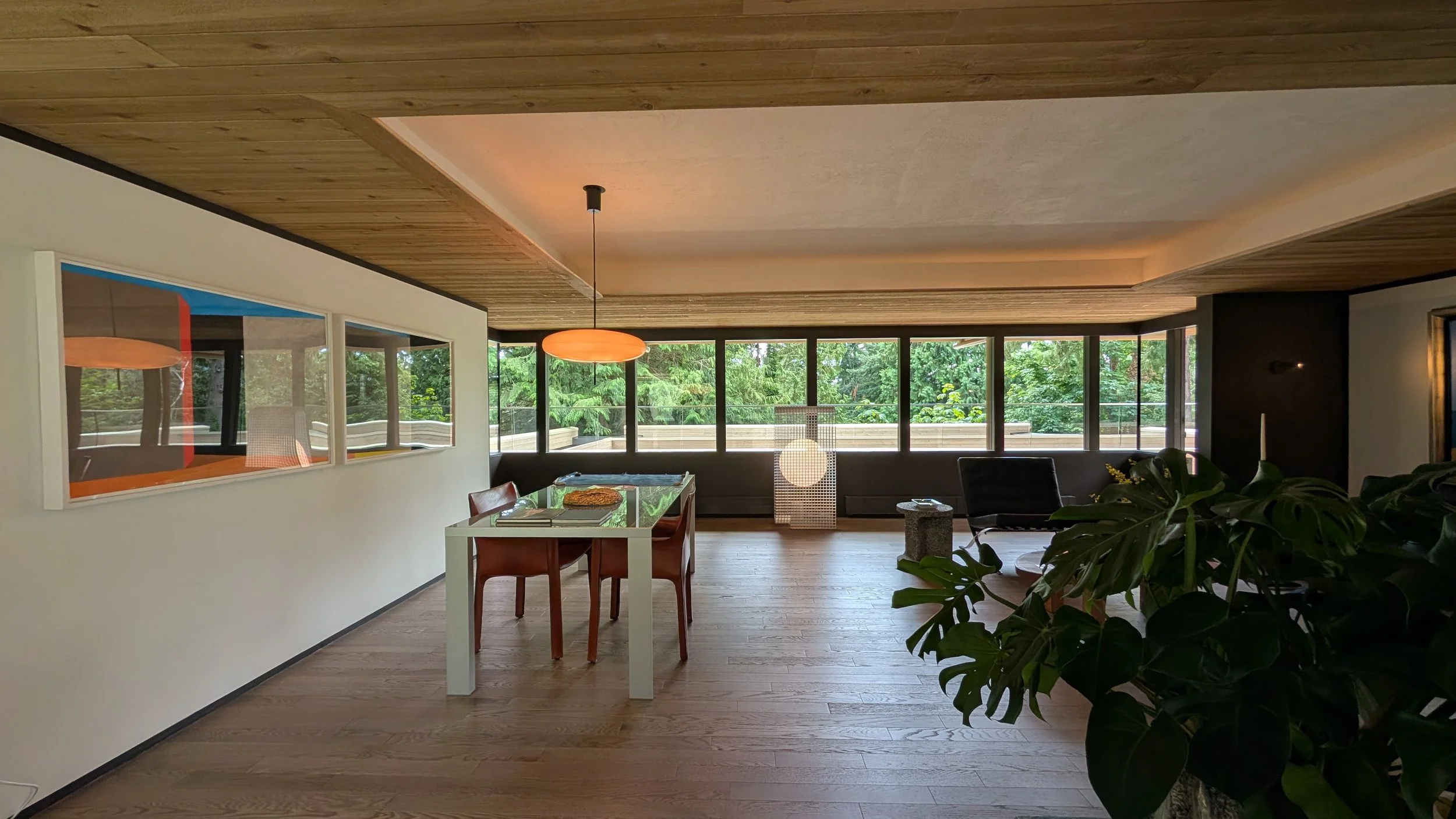Living room with large windows showing green trees outside, wooden ceiling and floor, a dining table with chairs, a black lounge chair, a potted plant, and a round pendant light.