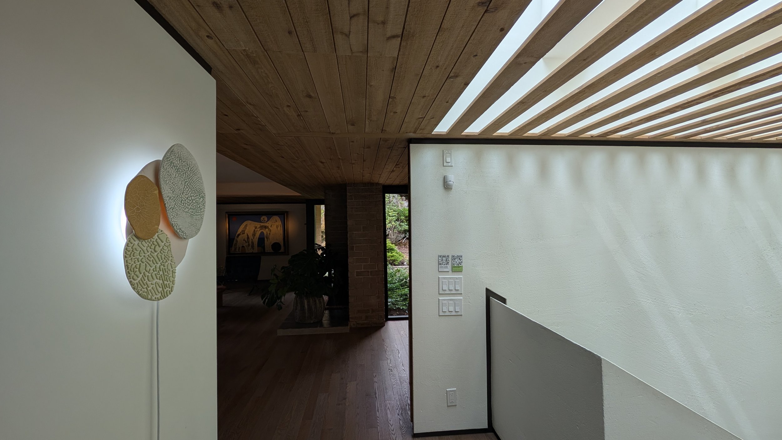 Interior view of a modern house with a wooden ceiling, white walls, and artwork on the wall. A large window lets natural light into the space.