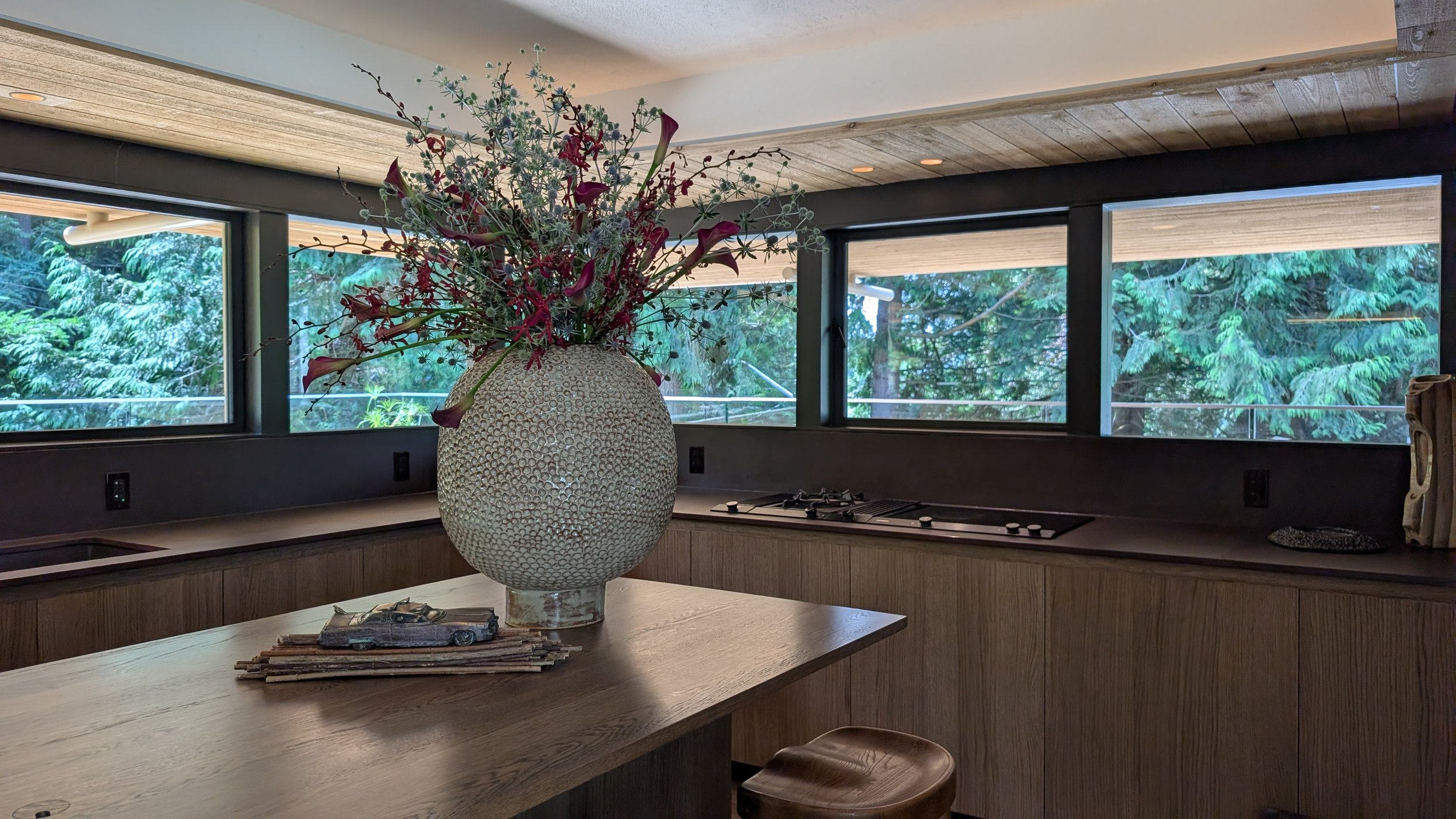 A modern kitchen with large windows showing green trees outside, a wooden table with a decorative vase of flowers, and kitchen appliances.