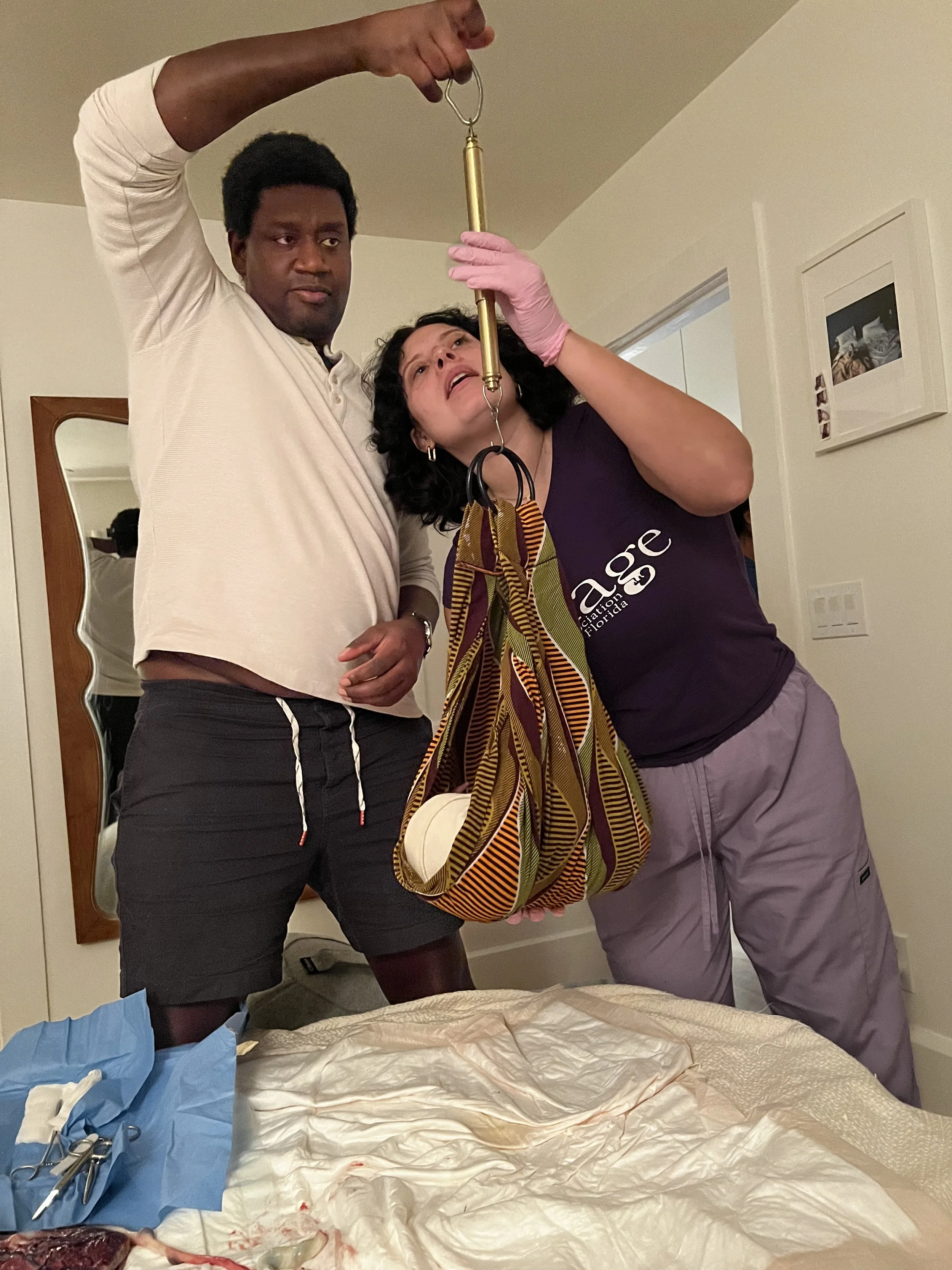 Two people perform a medical procedure, one holding a surgical tool and the other assisting, in a bedroom with medical supplies on the bed.