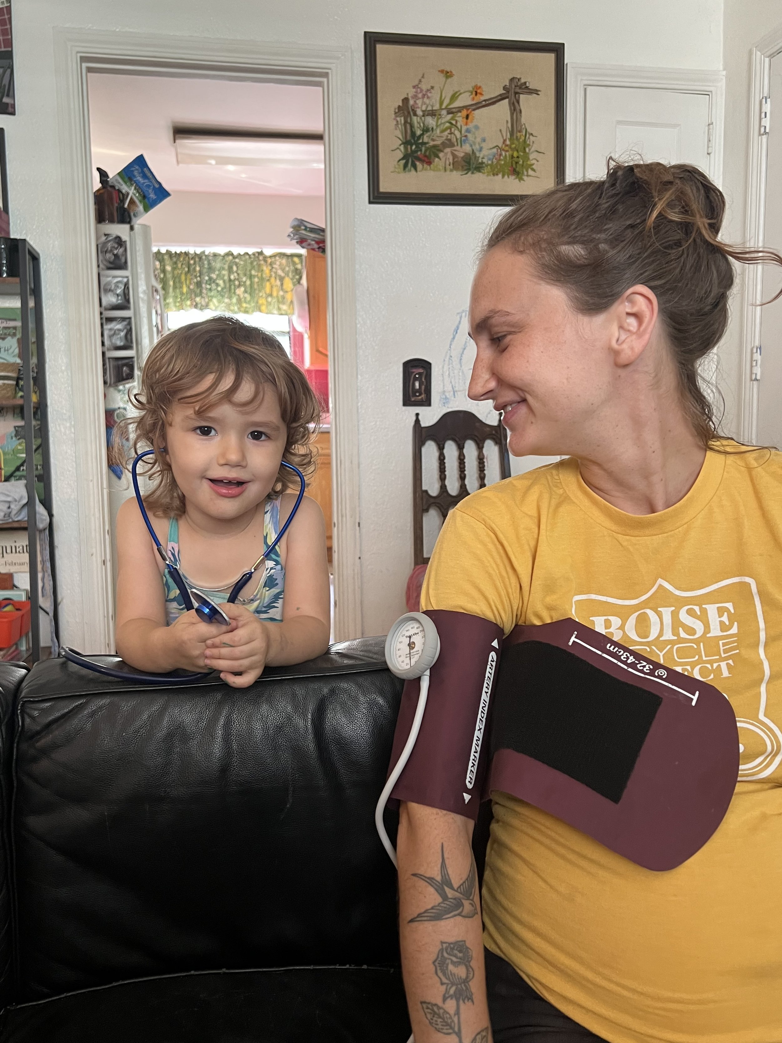 A young girl with a stethoscope around her neck talking to a woman with tattoos, who is having her blood pressure taken at home.