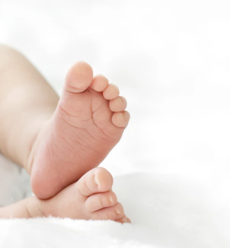A newborn baby lying on a soft blanket, wearing a white long-sleeve shirt, with one hand near their head, eyes closed.