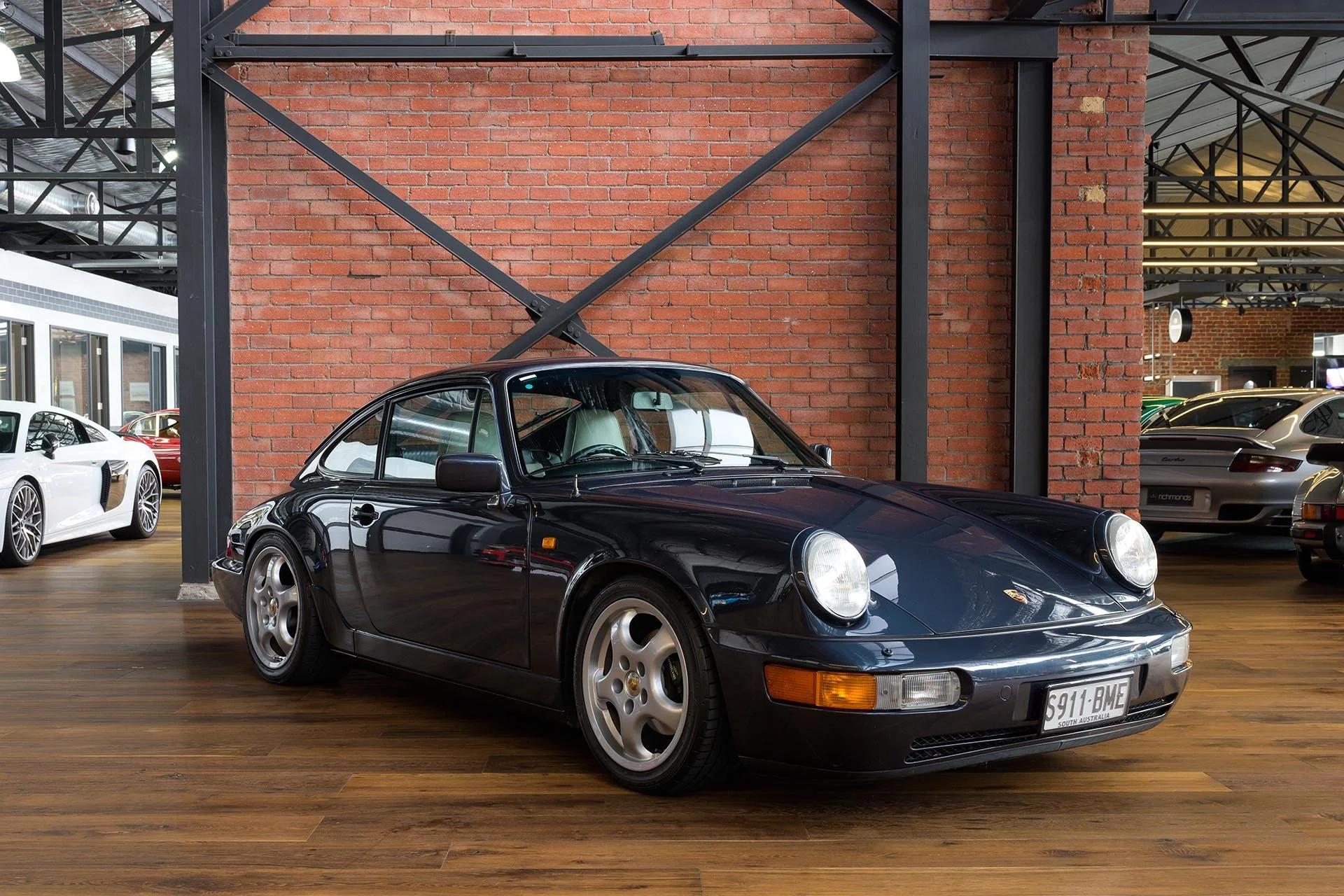 A black vintage Porsche 911 sports car inside a showroom with wooden floors and exposed brick walls, surrounded by other luxury cars.