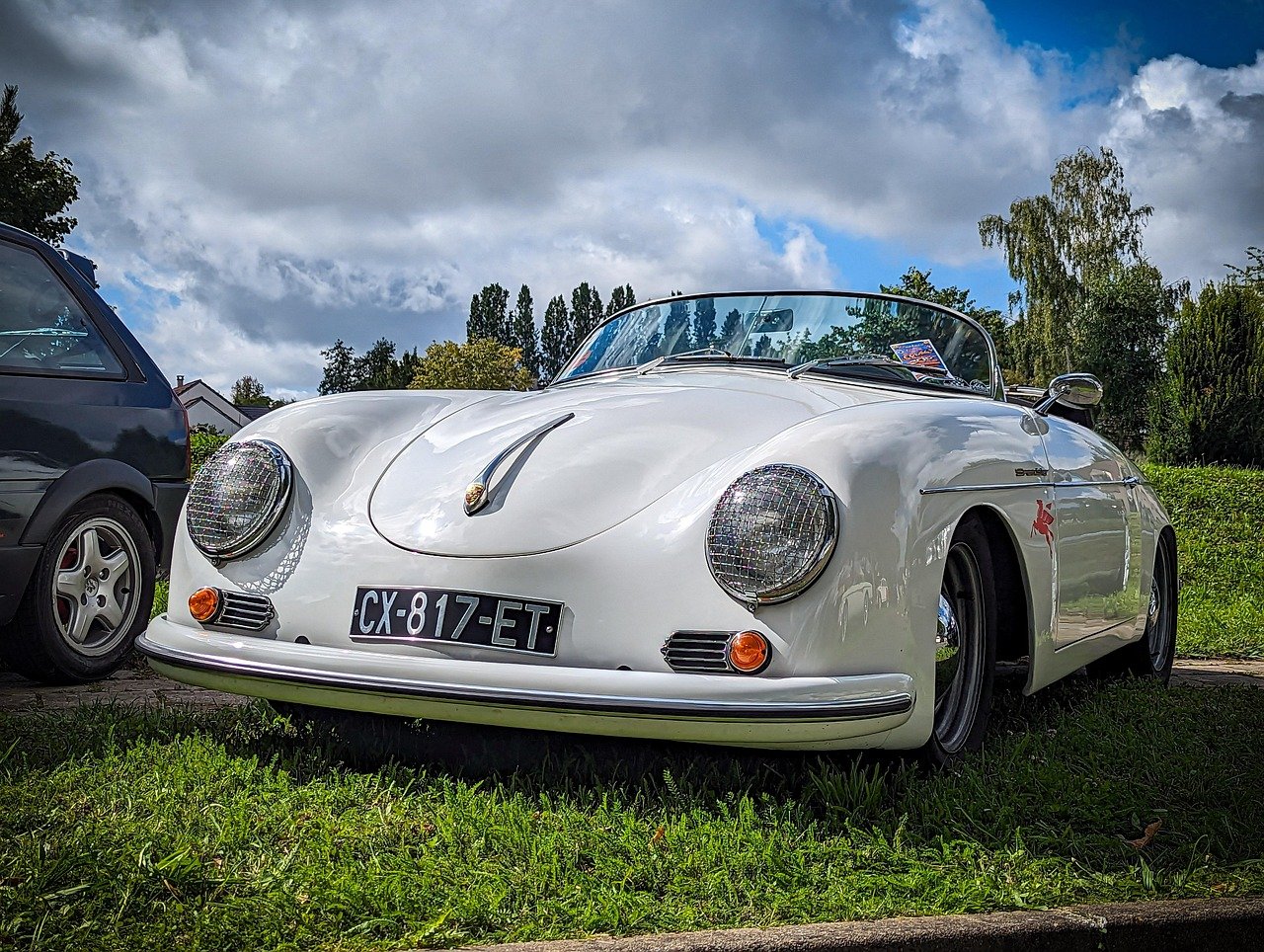 A white vintage Porsche convertible car parked on grass next to another vehicle, under a cloudy sky with trees in the background.