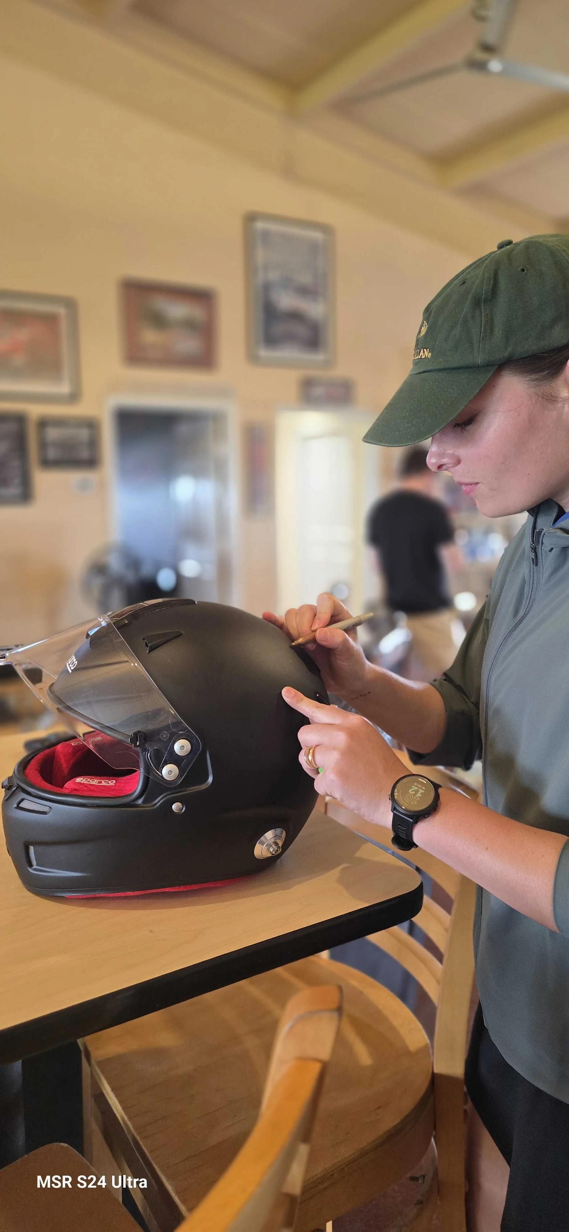 Lara from Longhorn Racing Academy signs a black Sparco Racing helmet owned by Alexander Wiener with a gold Sharpie on a wooden table inside the Harris Hill Raceway clubhouse.