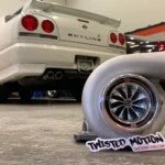 Close-up of a white sports car with visible rear lights and panther logo, with a turbocharger in the foreground.