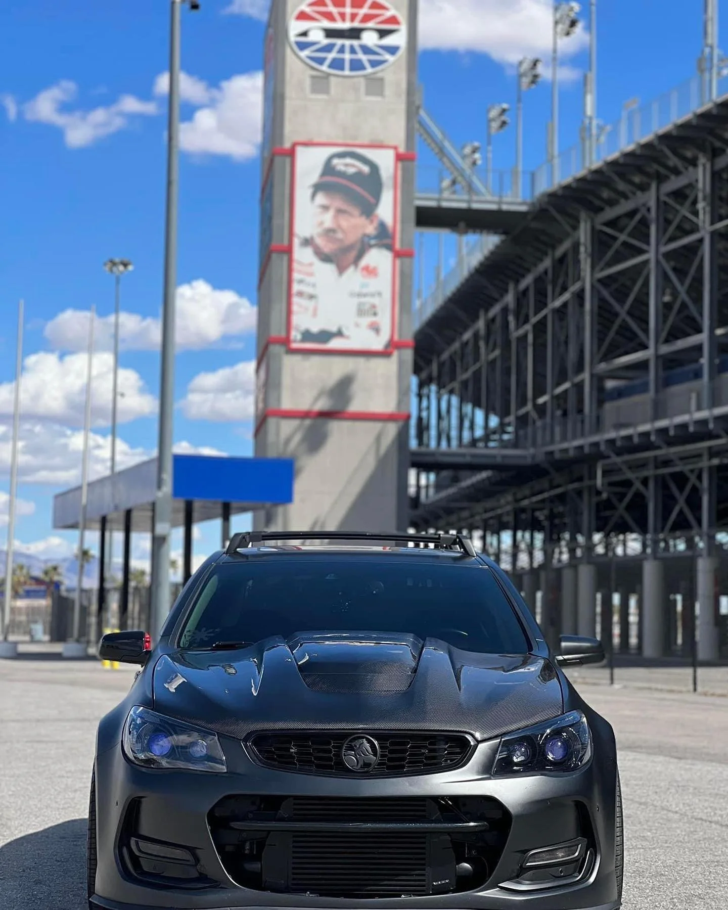 A black sports car parked in an empty lot with a racing stadium in the background, featuring stadium signage and a large billboard of a racing driver in front of a blue sky with scattered clouds.