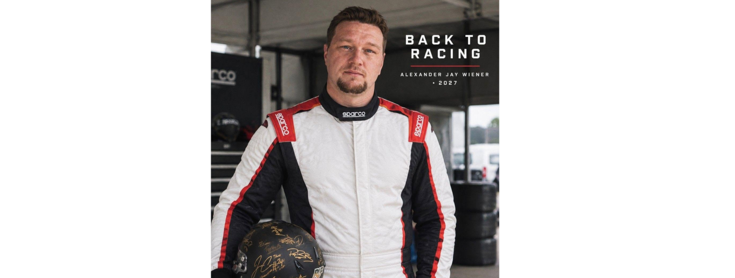 Team Owner Alexander (MSR) Wiener in a racing suit holding a black Sparco racing helmet with autographs stands in a garage or pit area, with tires and equipment in the background.