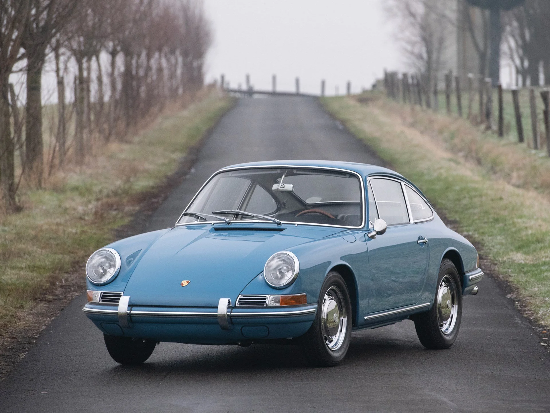 A vintage blue Porsche 911 car parked on a rural paved road with trees and a fence on either side, leading up a hill into foggy background.