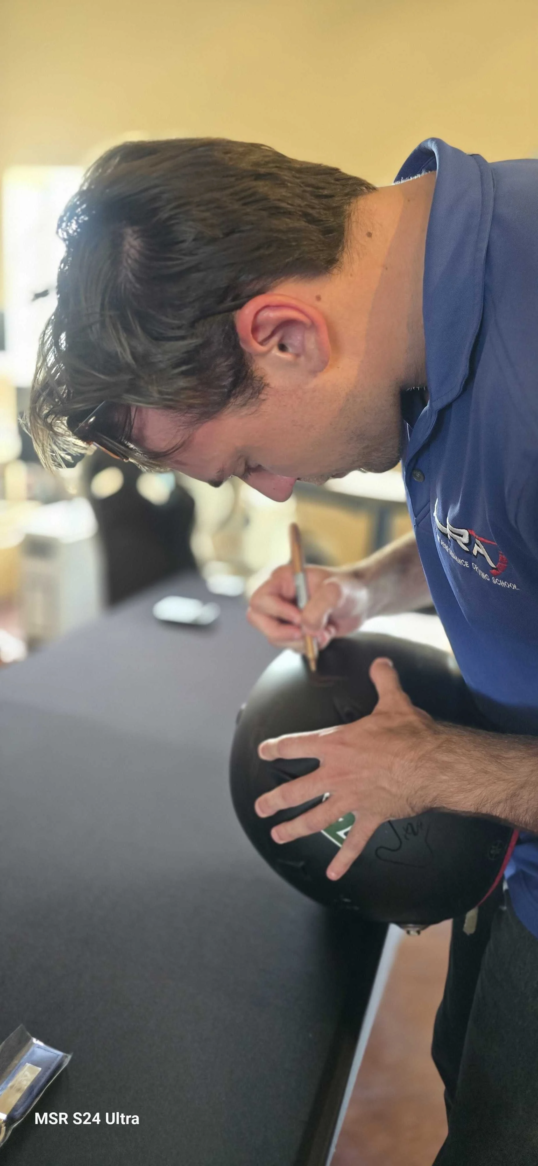 Another driver coach from Longhorn Racing Academy is signing a black Sparco helmet owned by Alexander Wiener with a gold Sharpie on a table inside the Harris Hill Raceway clubhouse.