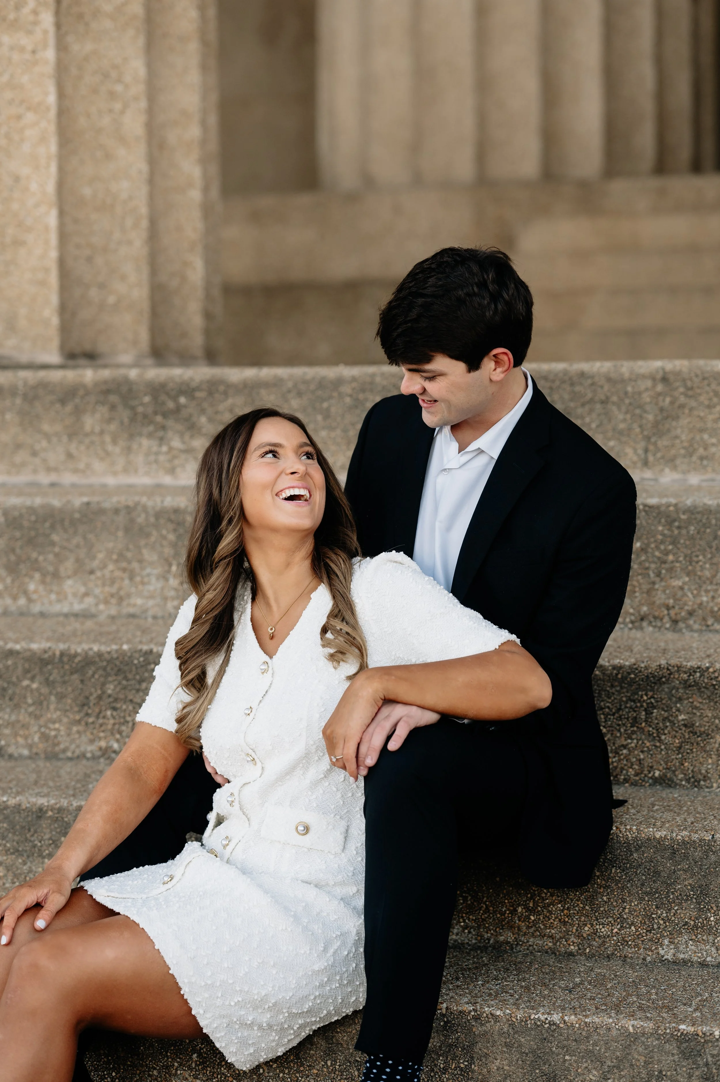A smiling couple sitting on steps outside, the woman in a white dress and the man in a black suit, sharing a joyful moment.