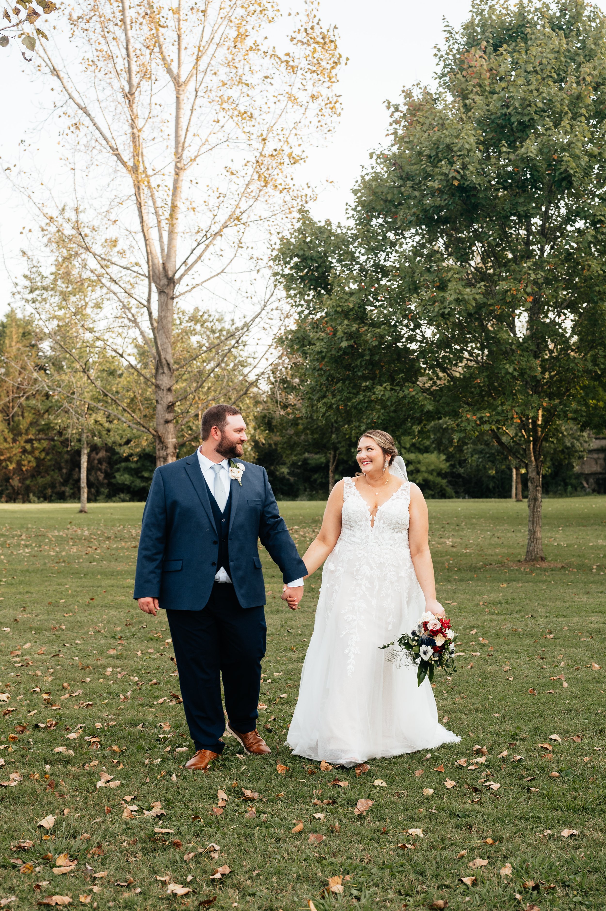A bride and groom walking hand in hand outdoors on a grassy field with trees in the background, smiling at each other during their wedding.