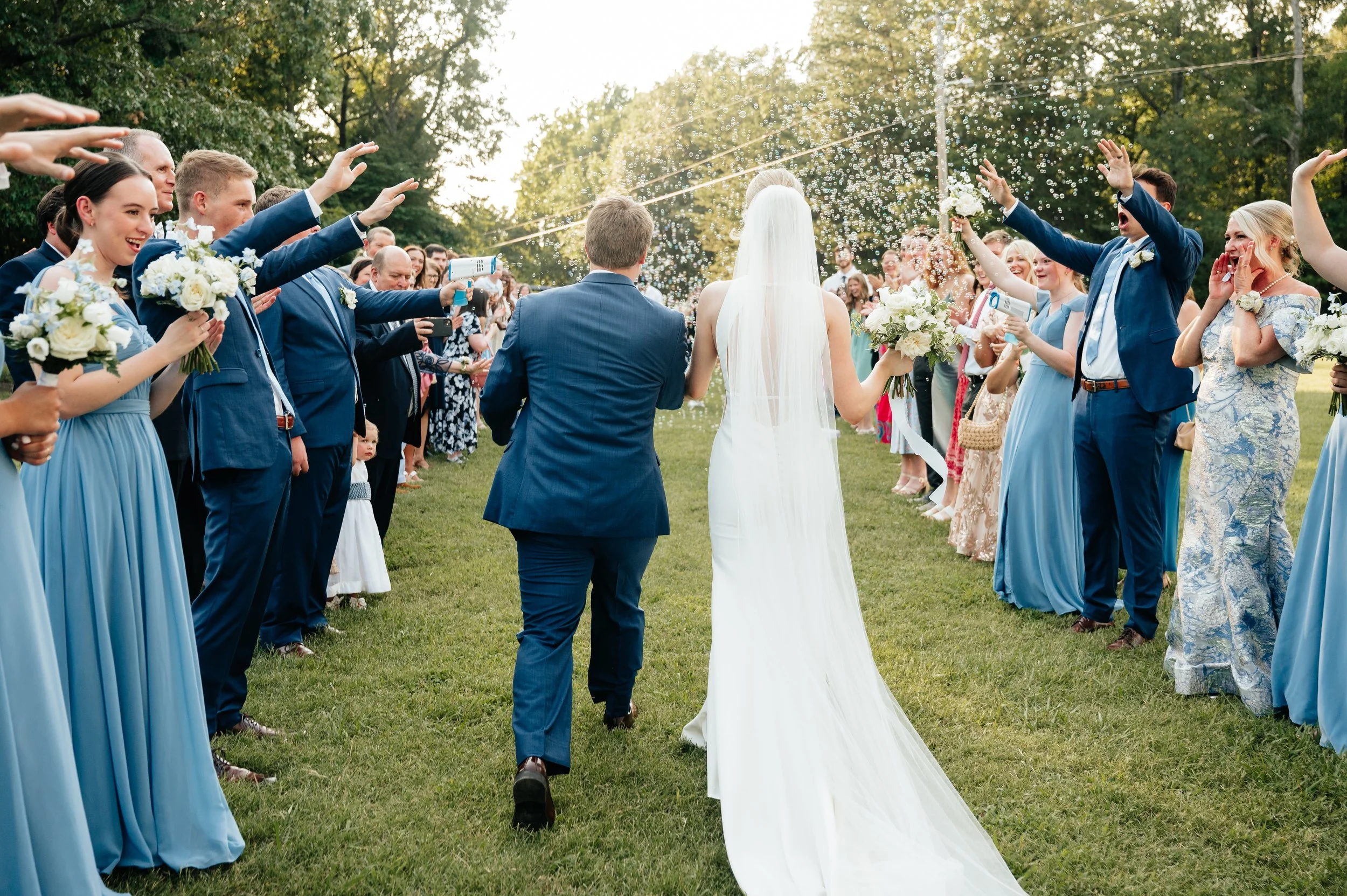 Bride in white wedding dress and veil holding bouquet walking with groom in blue suit, flanked by wedding guests in blue and floral dresses, celebrating outdoors on grass with trees in the background