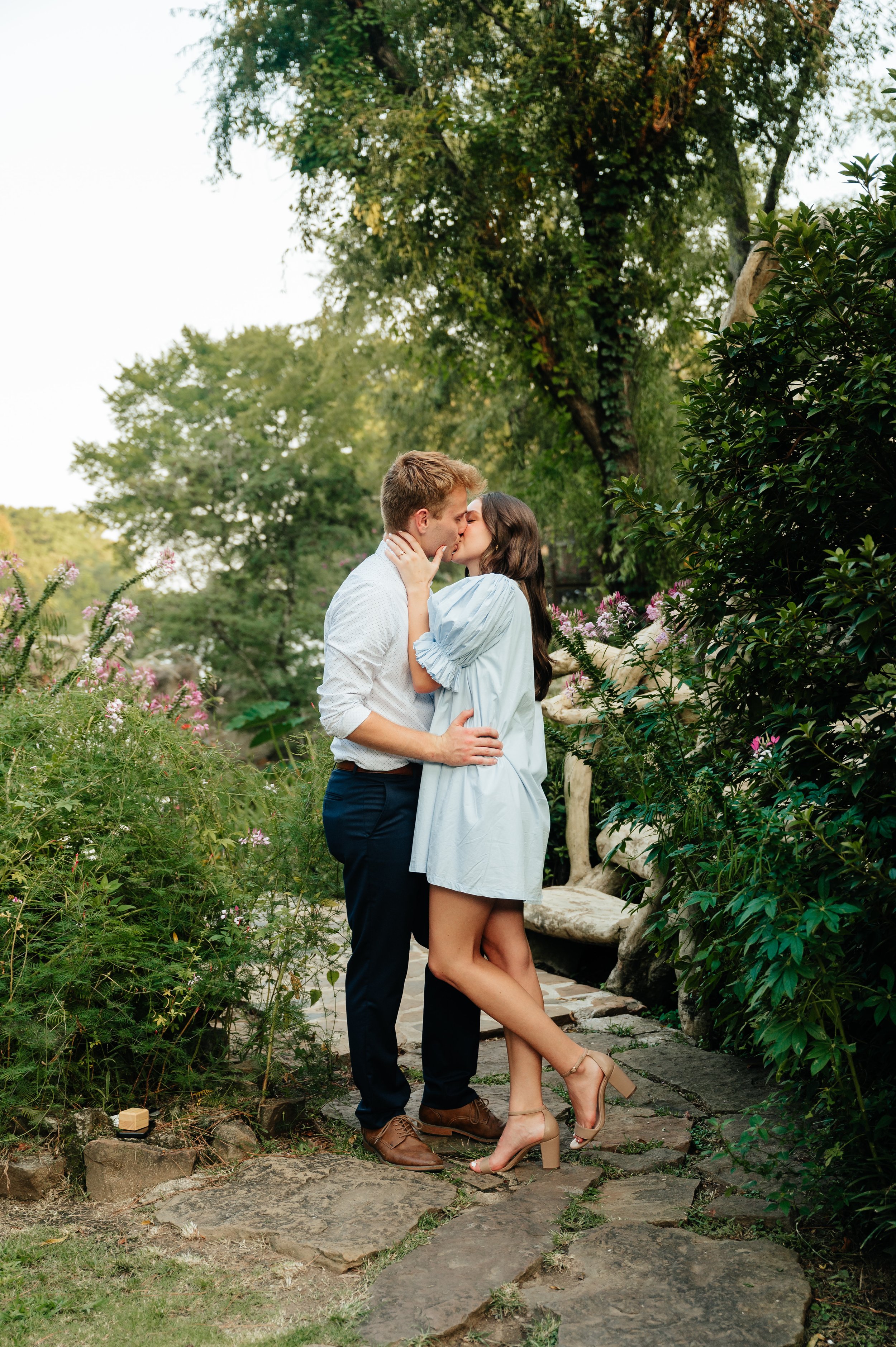 A couple kissing on a stone pathway in a lush garden surrounded by greenery and flowers.