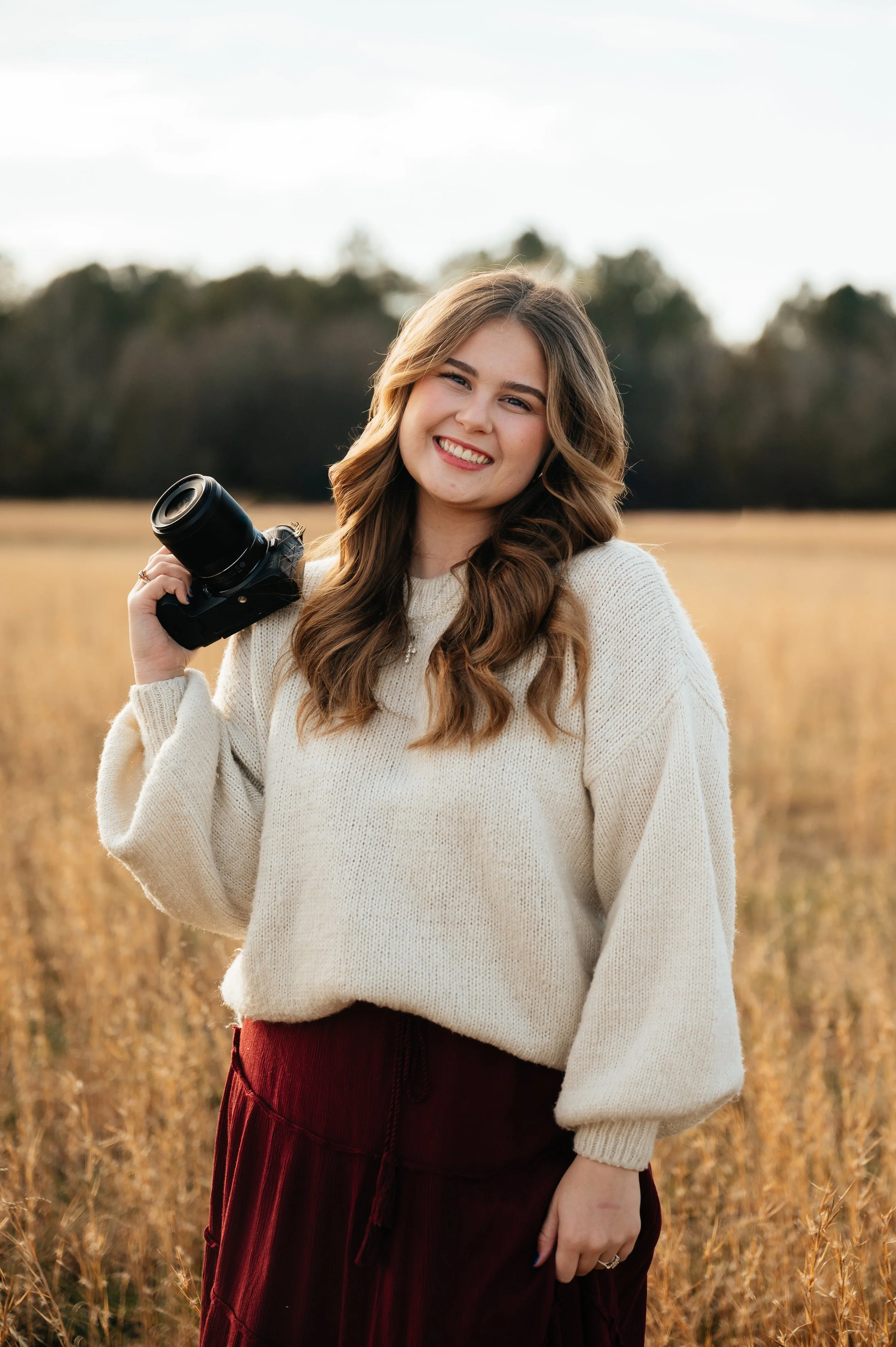 A young woman with long brown hair, smiling, holding a camera in a field with tall grass, dressed in a cream sweater and maroon pants, during daylight.