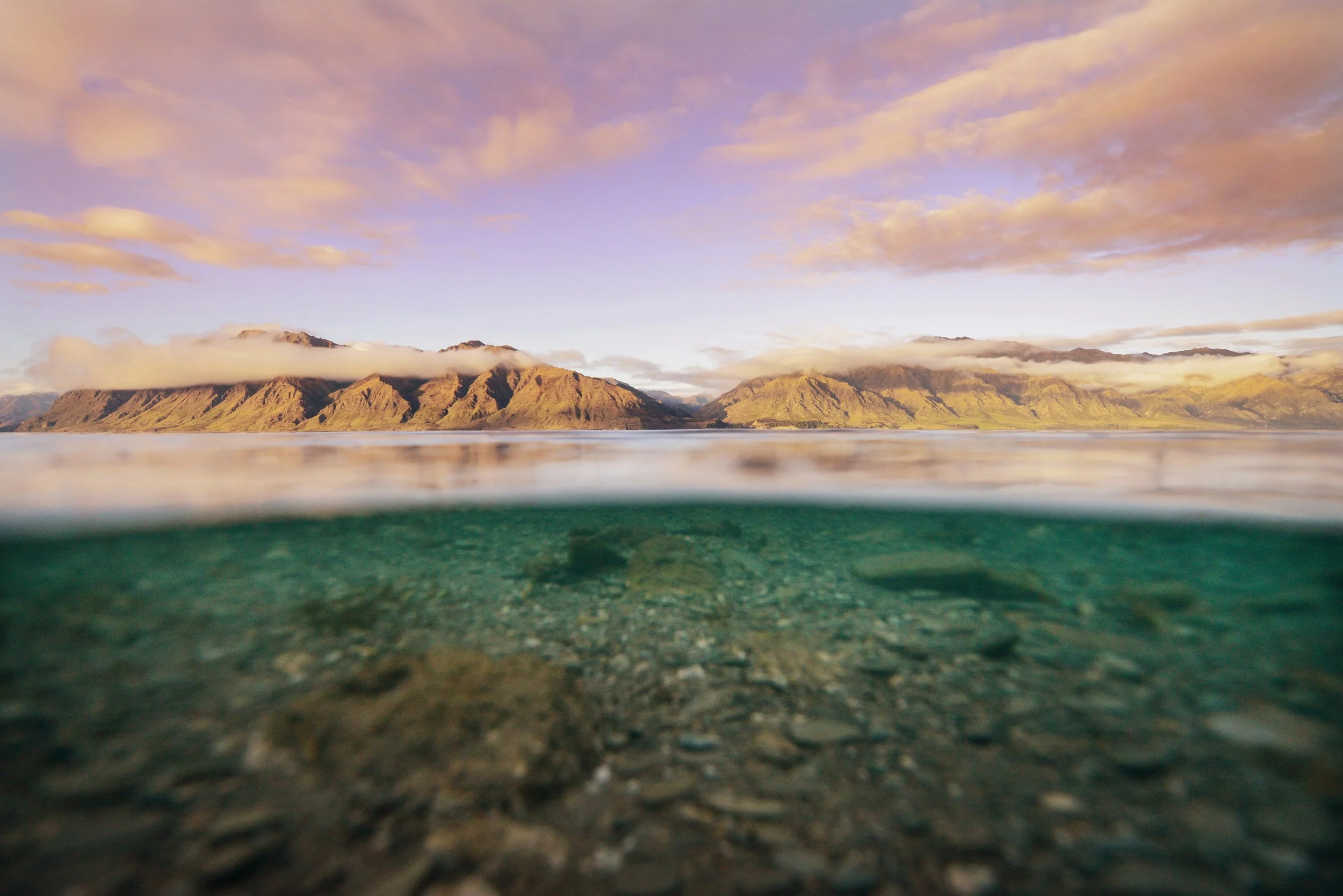 A scenic view of mountains in the distance with a lake in the foreground, half submerged, showing rocks beneath the water's surface during sunrise or sunset.