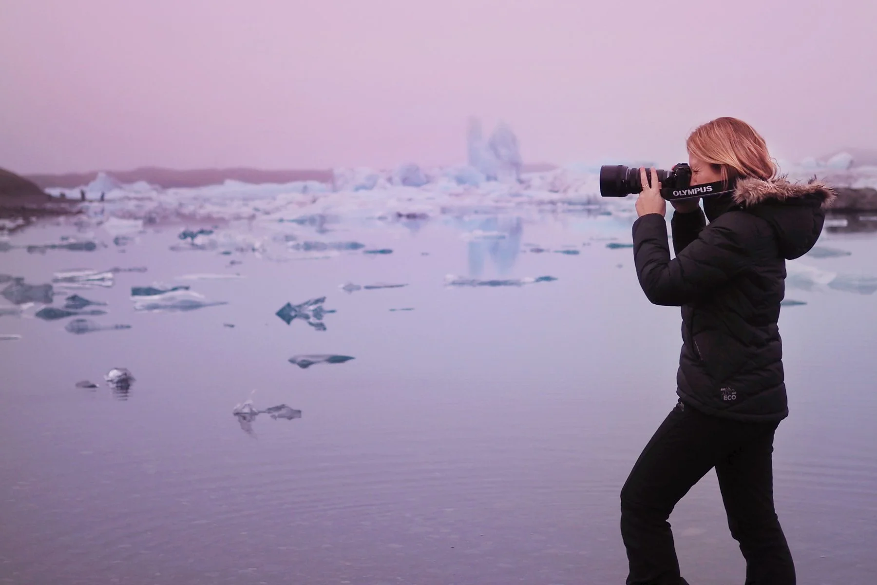 A female photographer with blonde hair wearing a black winter coat taking photos with a camera in a cold, icy landscape with water and floating ice chunks, pink and purple sky in the background.