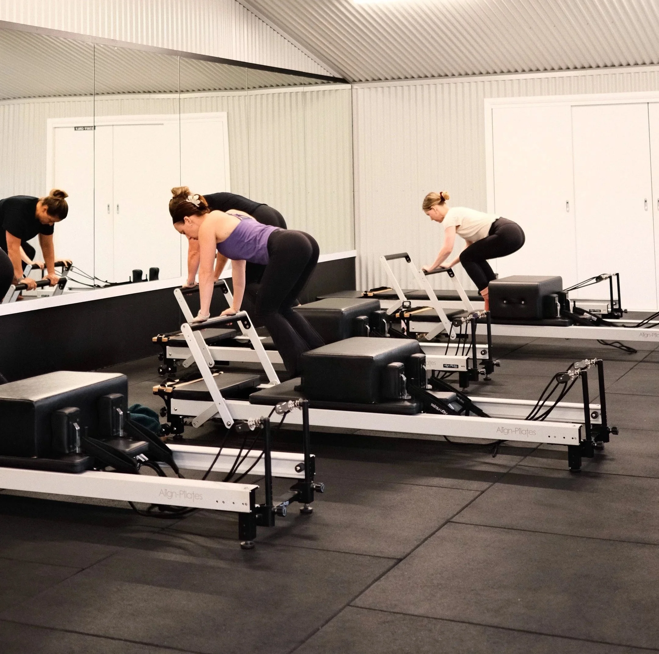 Four women in workout attire performing balancing exercises with pink dumbbells on reformer pilates machines in a fitness studio.