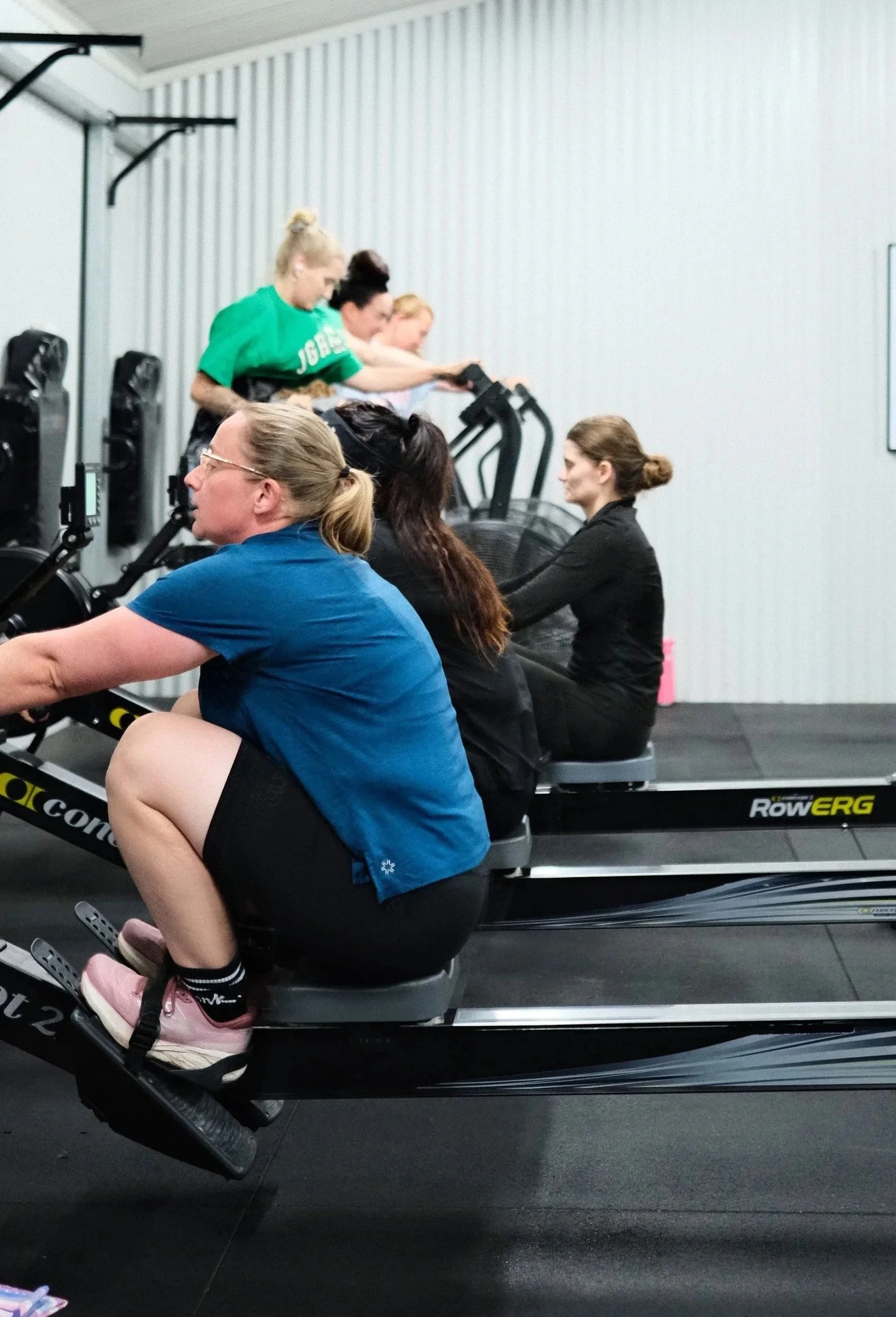 People exercising in a fitness gym, using rowing machines, kettlebells, and other equipment, with a high ceiling and large fans for ventilation.