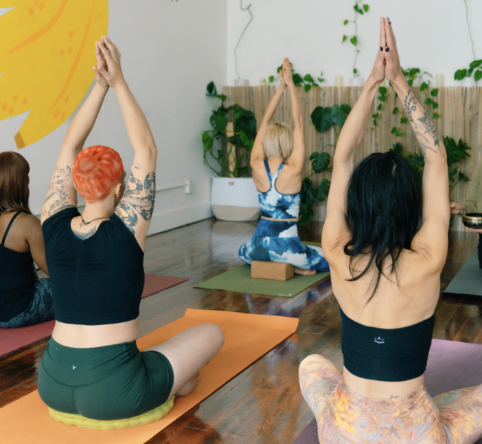 A group of beginner students practicing gentle yoga. They are seated with their legs crossed with their hands pressed over their heads.