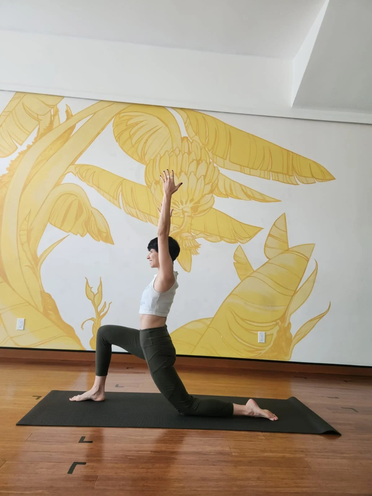 woman with short pixie haircut wearing cropped white tank tops and olive green leggings in a lowered lunge pose