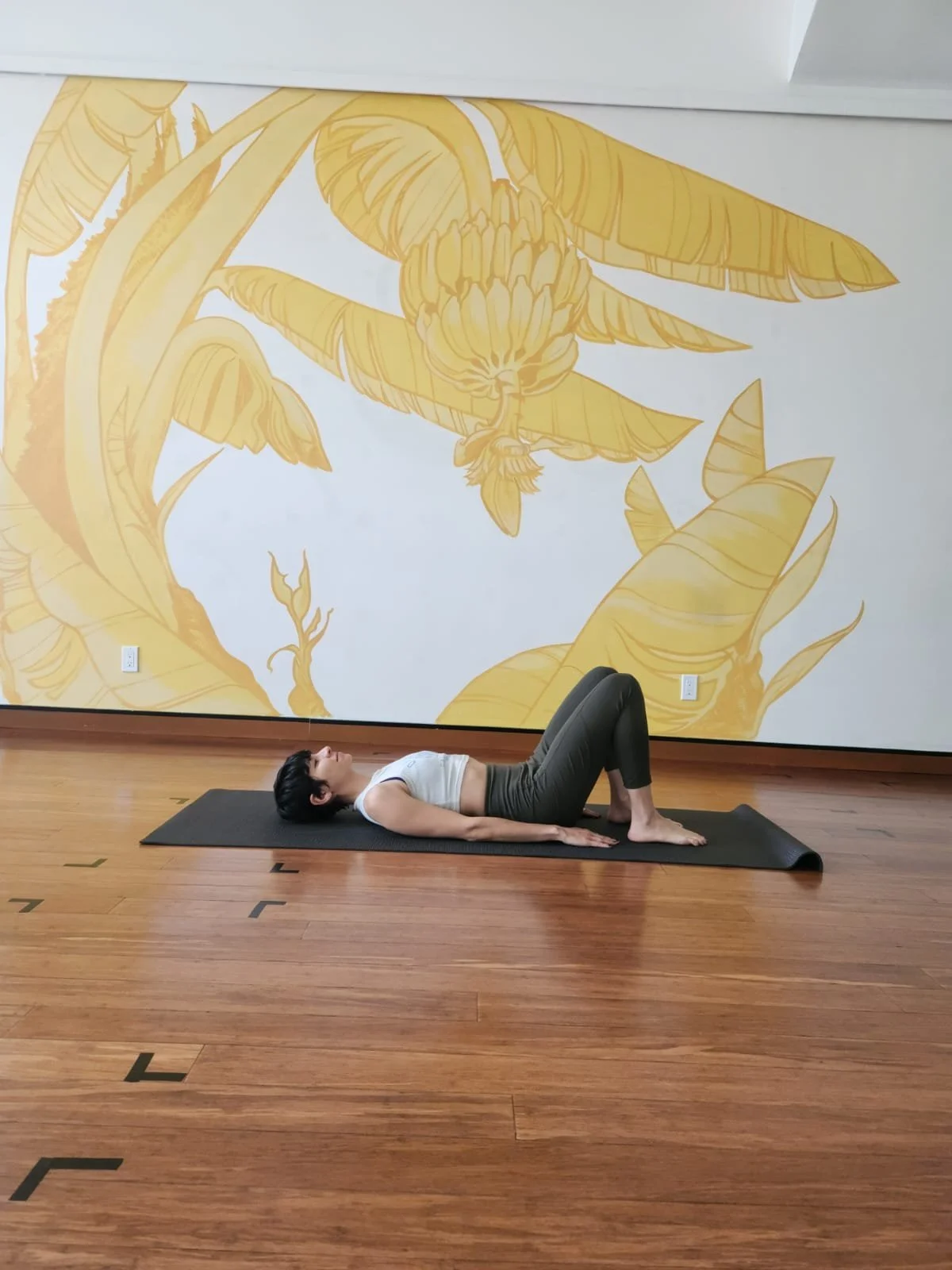 woman with short pixie haircut wearing a white cropped tank top and olive green leggings is laying on a yoga mat with her feet on the mat so that her knees are pointed up. Her arms are by her side.
