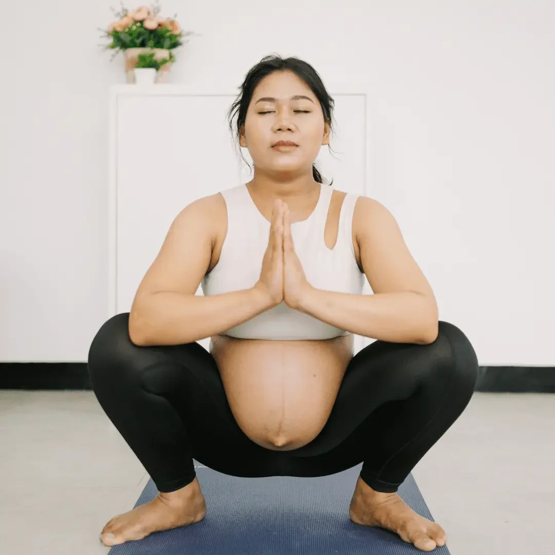 Asian woman with hair pulled back in a loose pony-tail wearing a white cropped yoga top and black legginds with her pregnant stomach exposes. She is doing malasana pose (yogi squat)