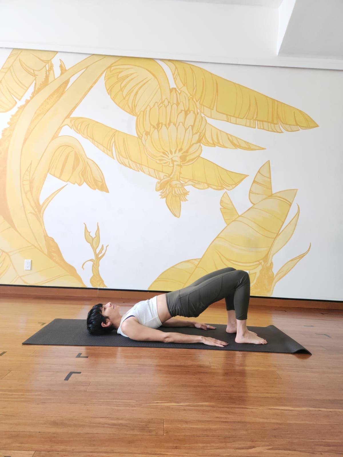 woman with short pixie haircut wearing a white cropped tank top and olive green leggings is laying on a yoga mat with her feet on the mat so that her knees are pointed up. Her arms are by her side and her hips are lifted off of the mat.